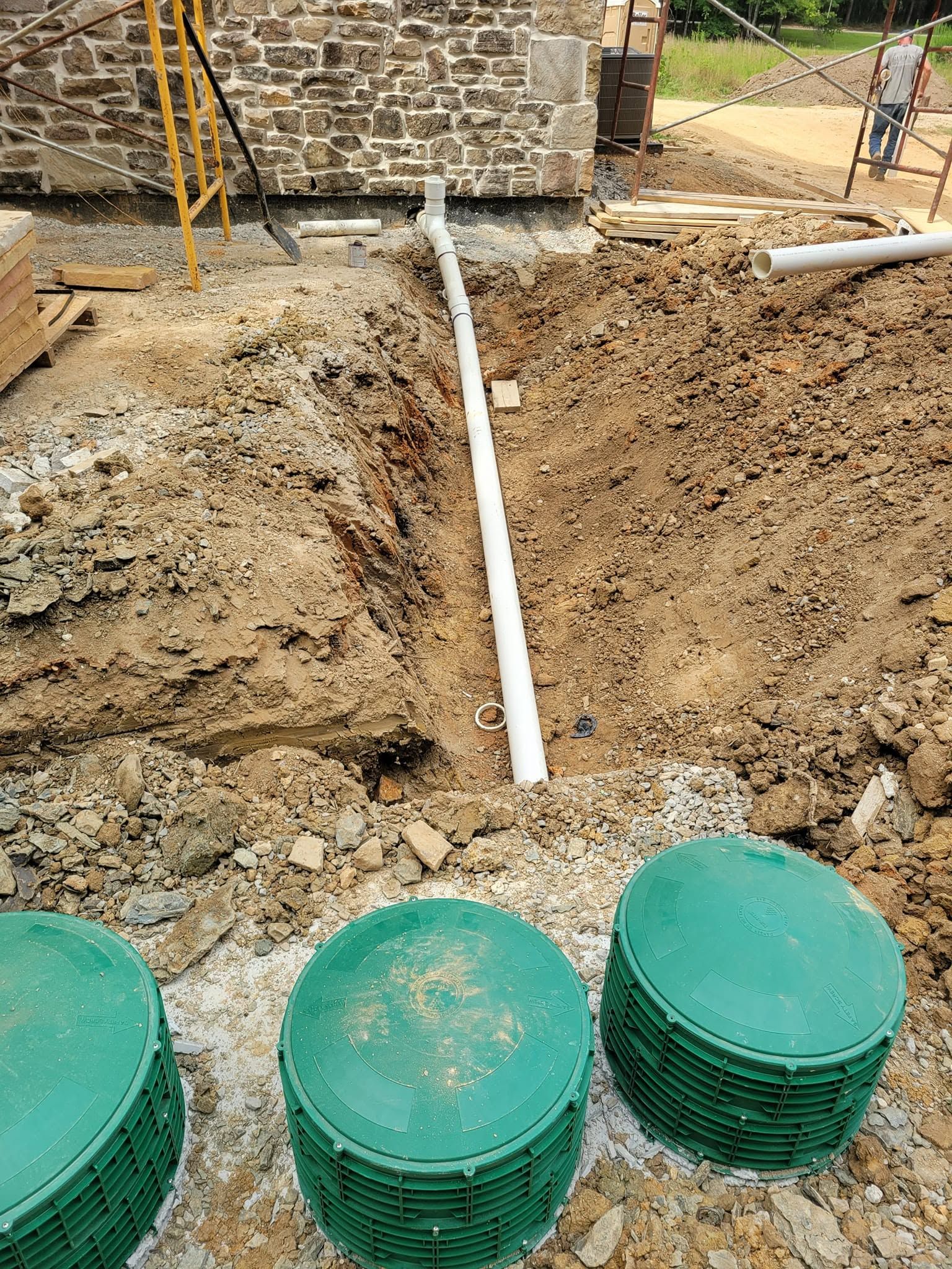 Construction trench with a white PVC pipe, dirt piles, and green pipe caps in the foreground.
