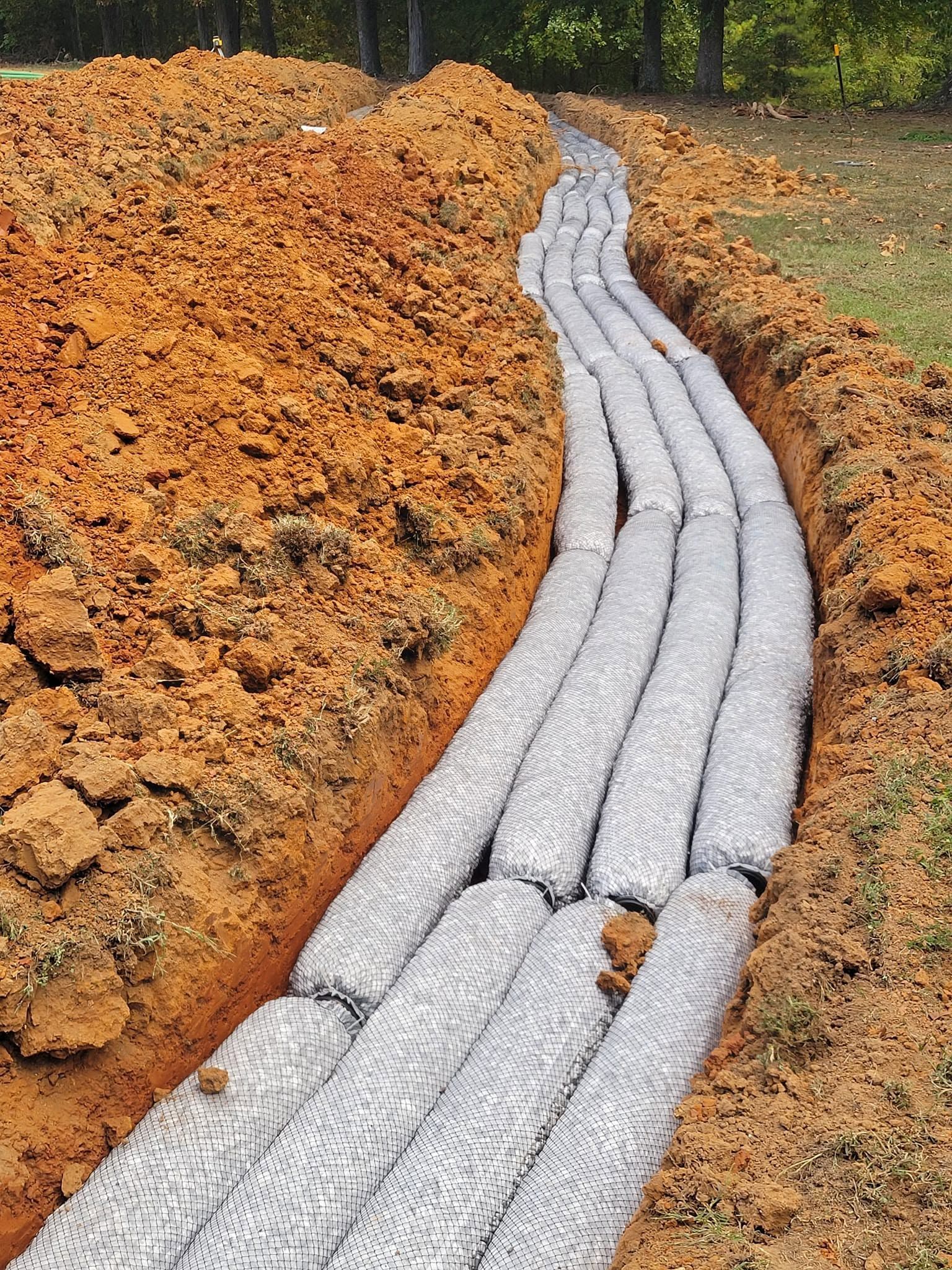 Black drainage pipes curve through an orange-brown earthen trench on a wooded slope.