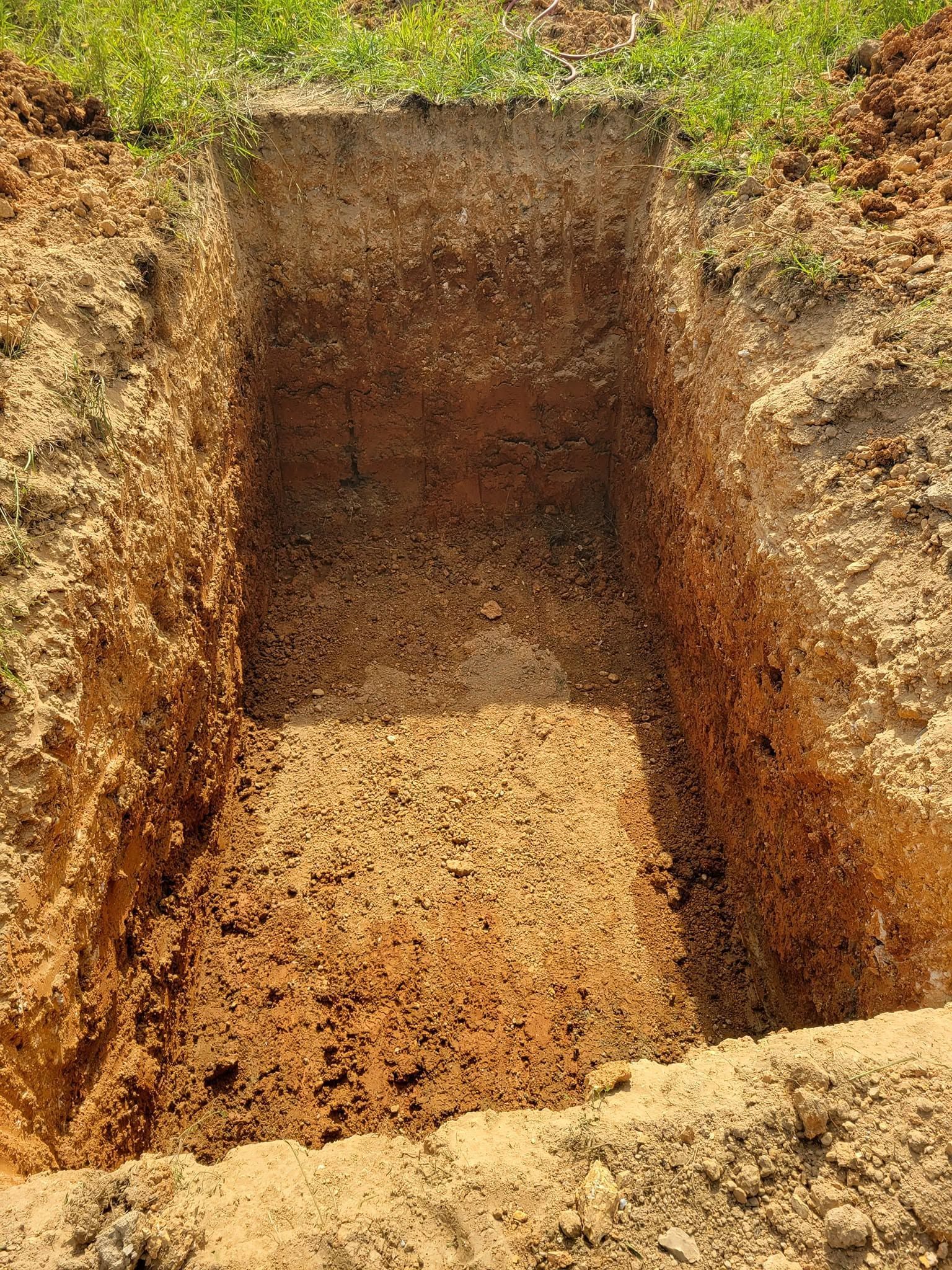 Empty rectangular pit in dry soil with grassy edges and exposed dirt walls