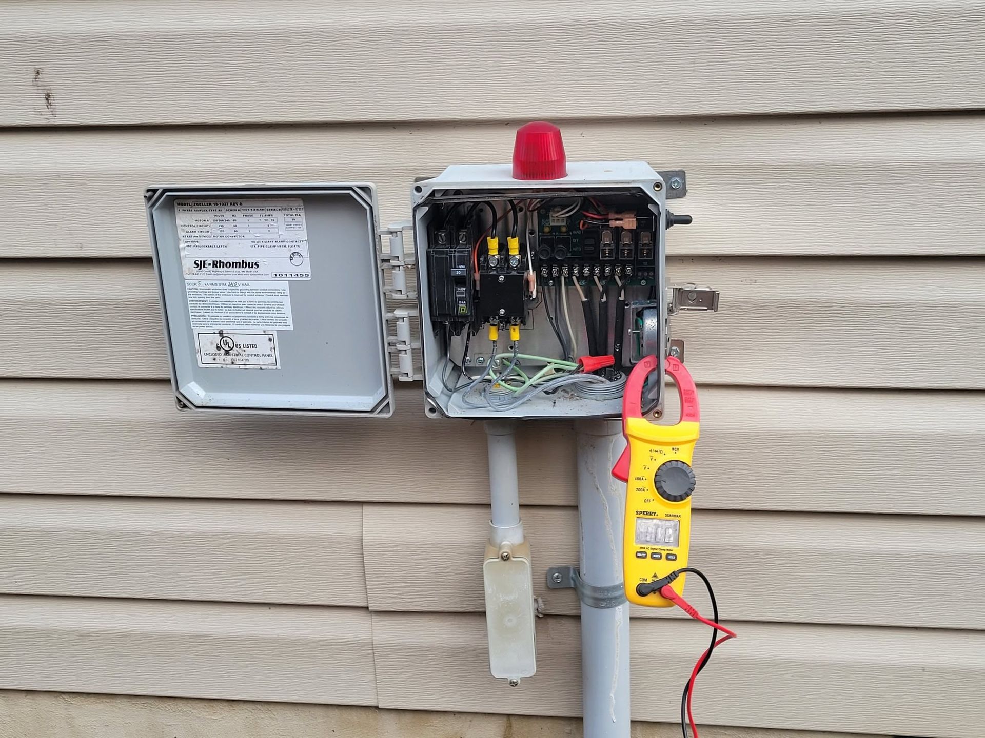 Open electrical panel on house exterior with red warning light and yellow multimeter clipped beside it