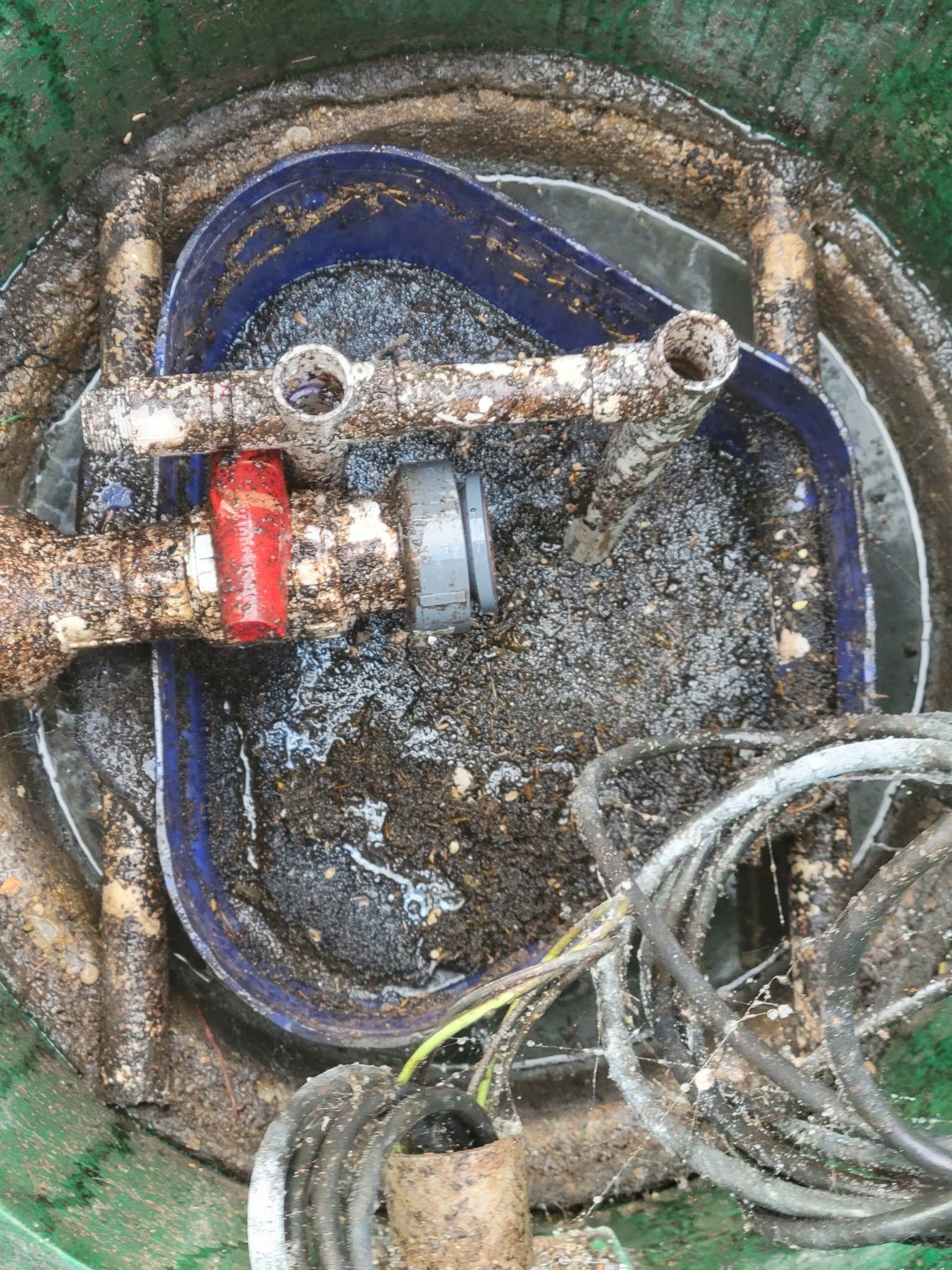 Close-up of a muddy utility valve box with blue piping, a red handle, and tangled cables.