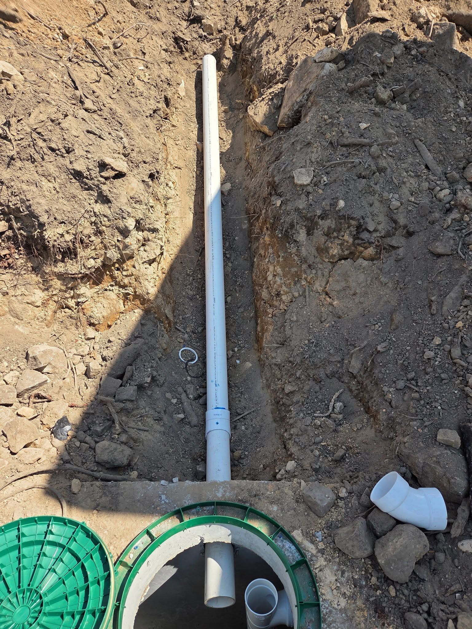 White pipe laid in a dirt trench beside a green lid and small fitting pieces.