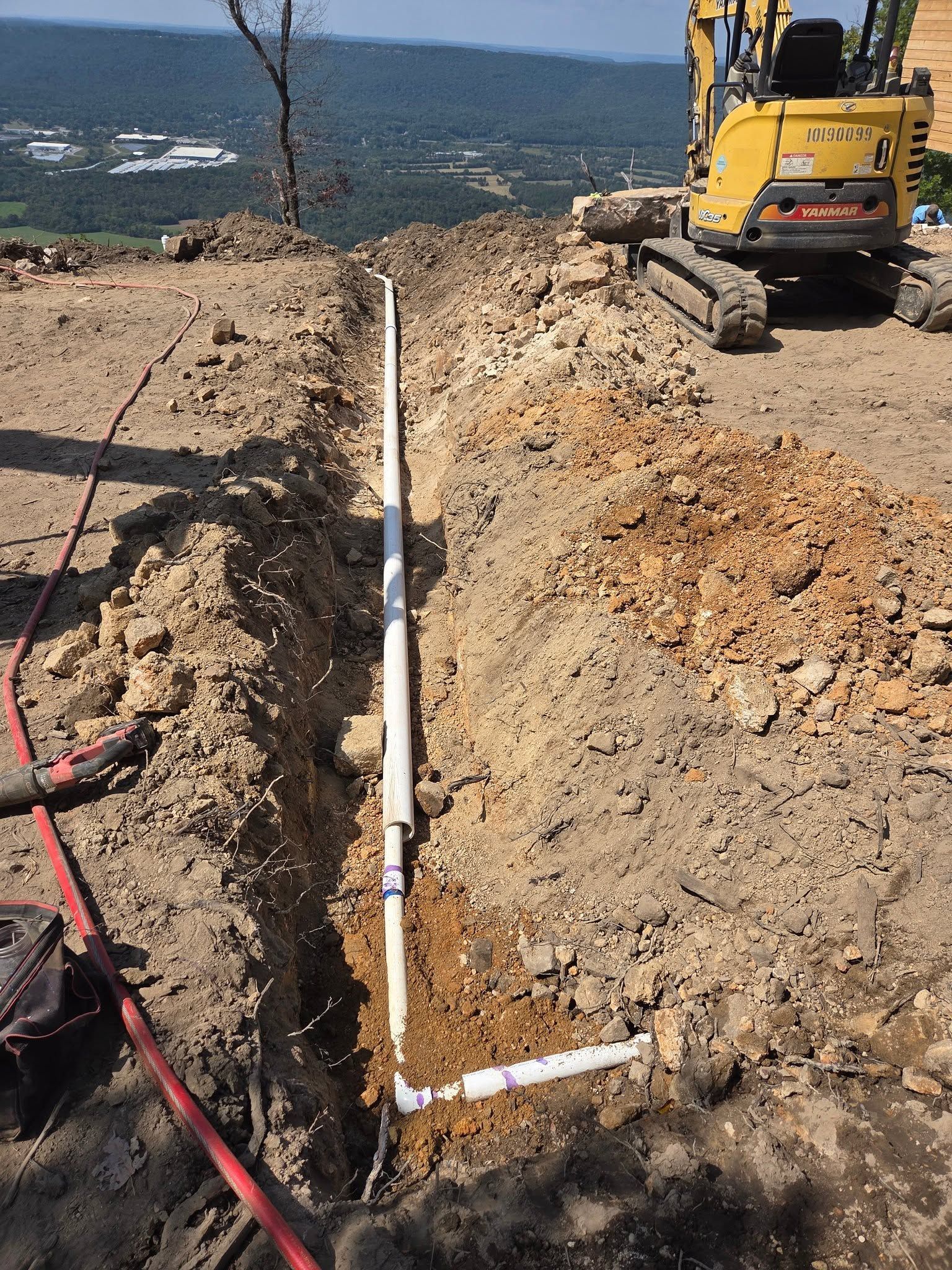 Excavation trench with white PVC pipe and yellow excavator beside a waterfront.