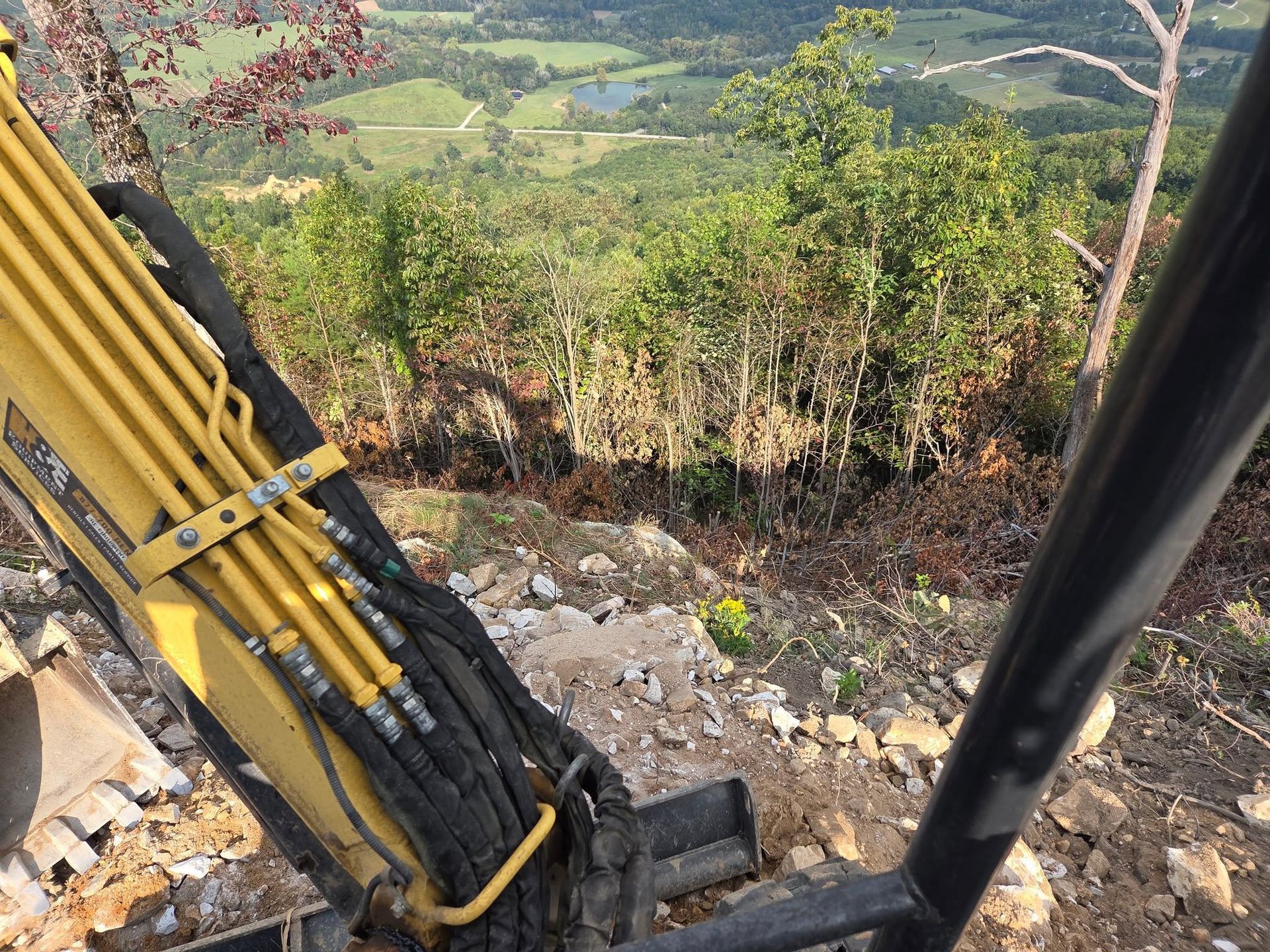Excavator arm on a rocky hillside overlooking a green valley and trees