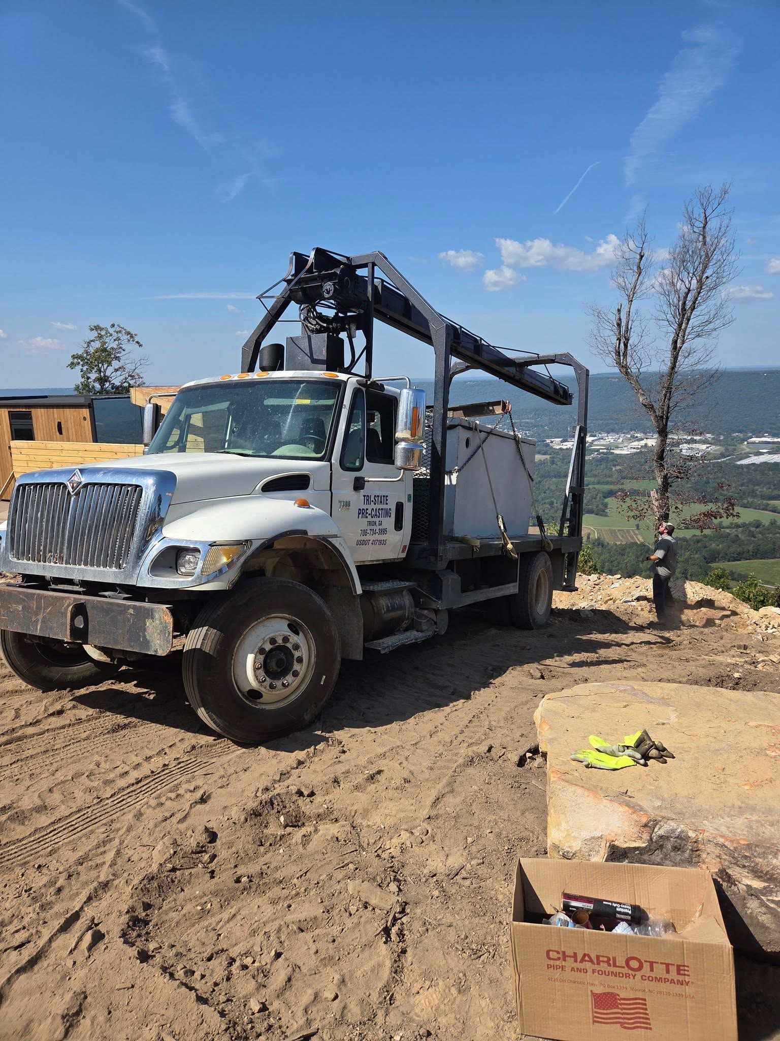 White utility truck with crane parked on dirt beside rocks and a leafless tree under a blue sky.