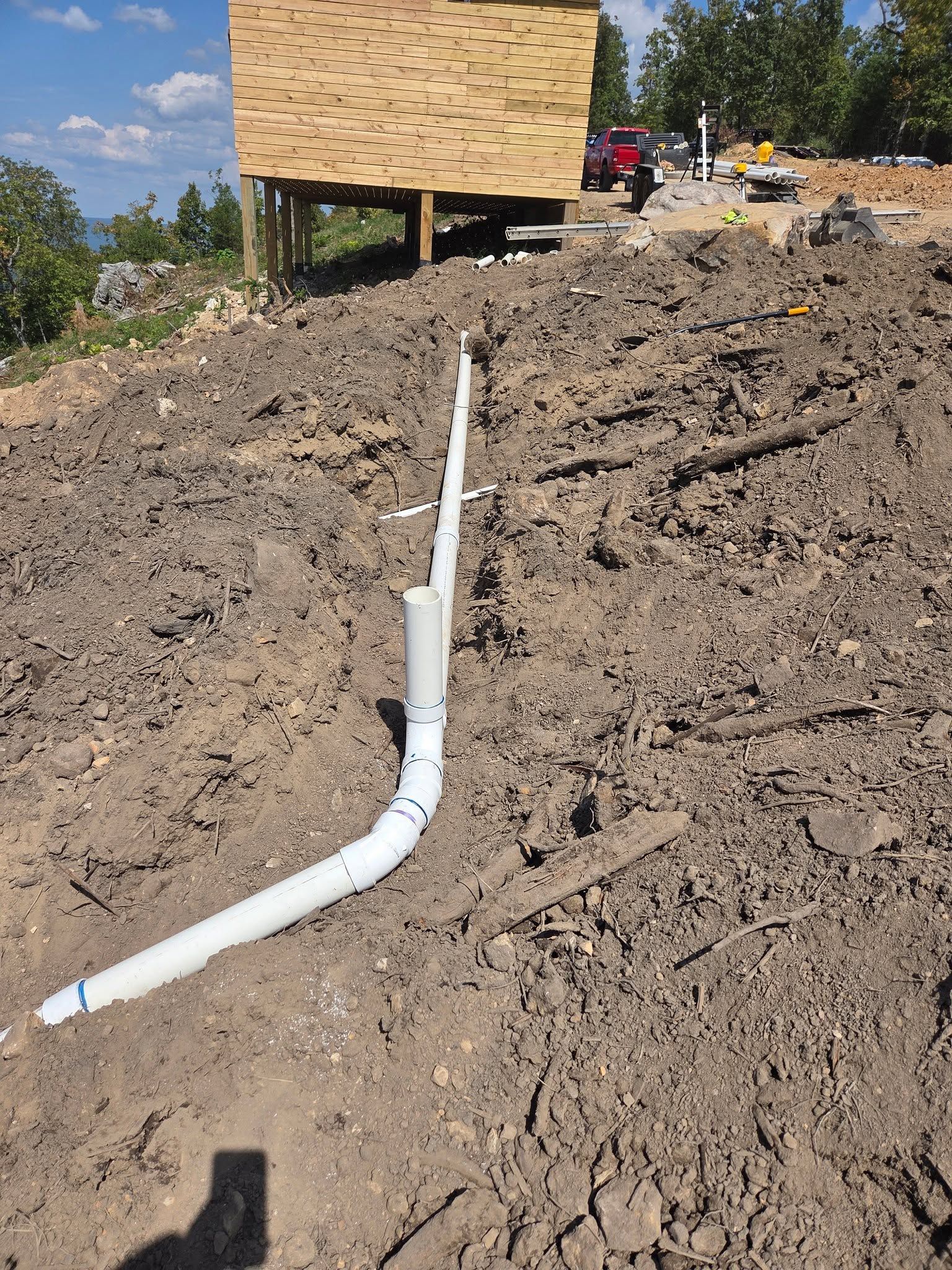 White drainage pipe runs up a dirt slope toward a wooden building at a construction site.