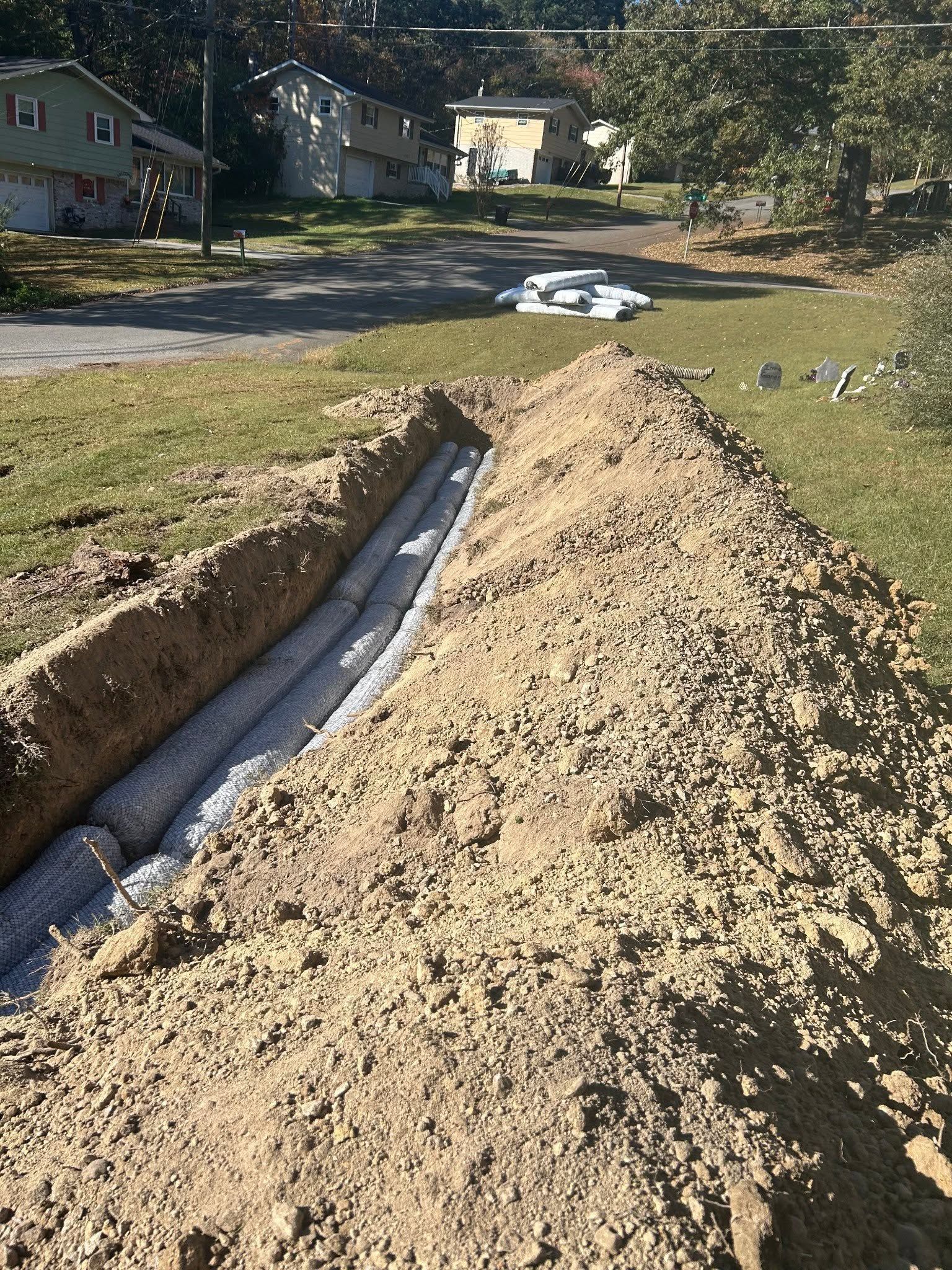 Trenched yard with a black drainage pipe and large mound of dirt in a suburban neighborhood.