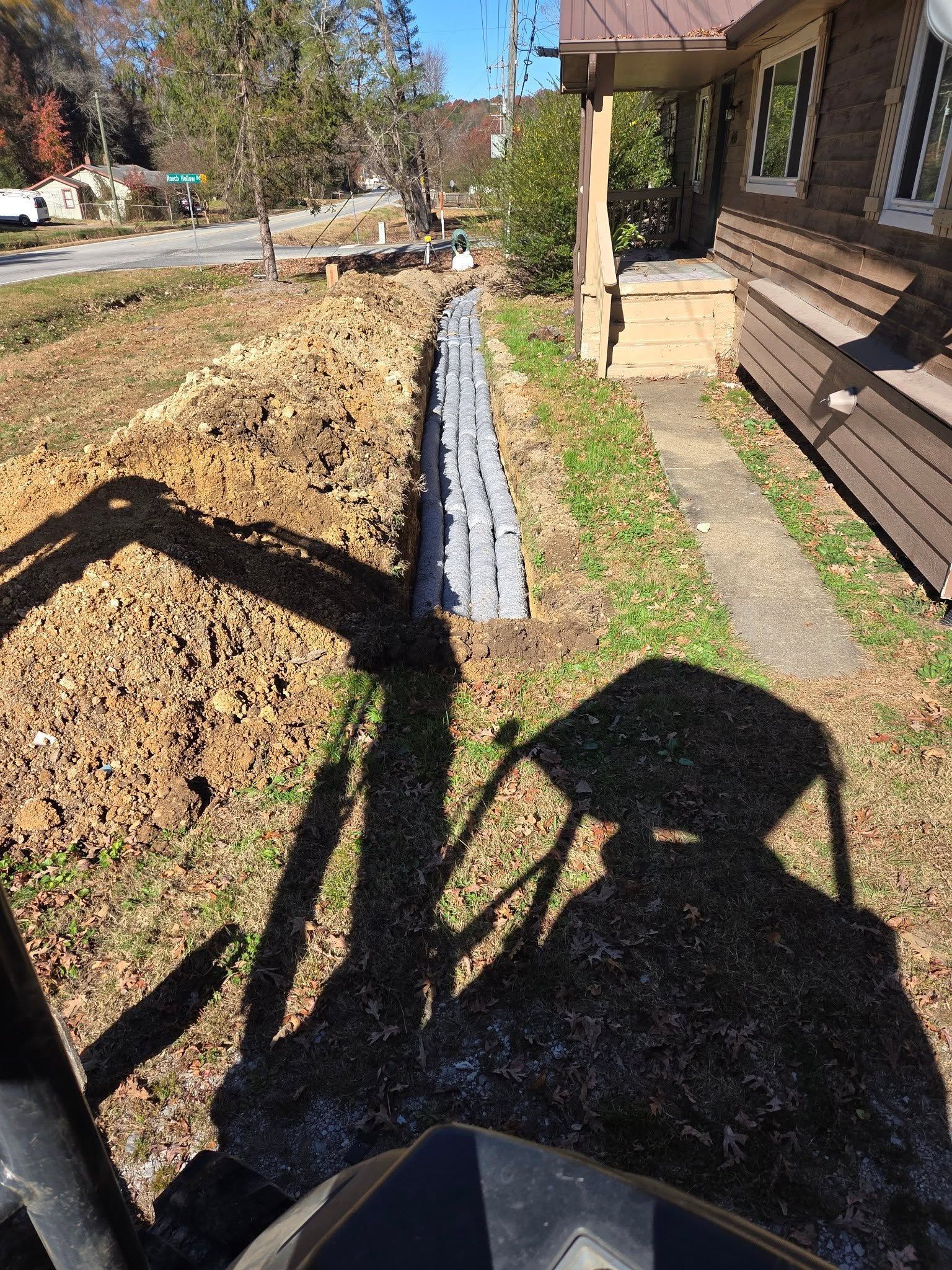 Front yard beside a house with a black edging strip along a dirt garden bed and a person’s shadow on the grass