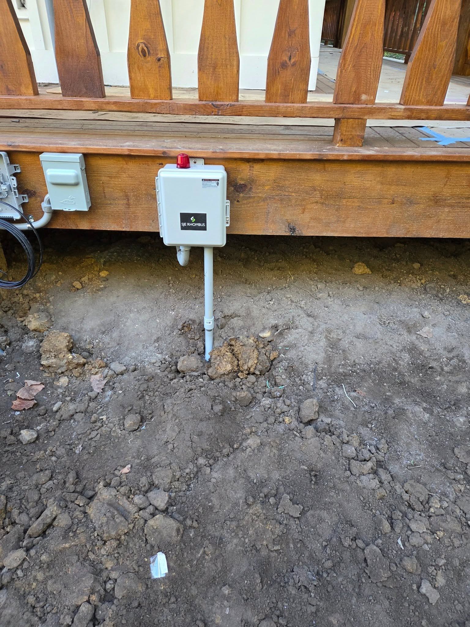 Small white electrical box mounted on a wooden fence above bare dirt, with conduit running downward