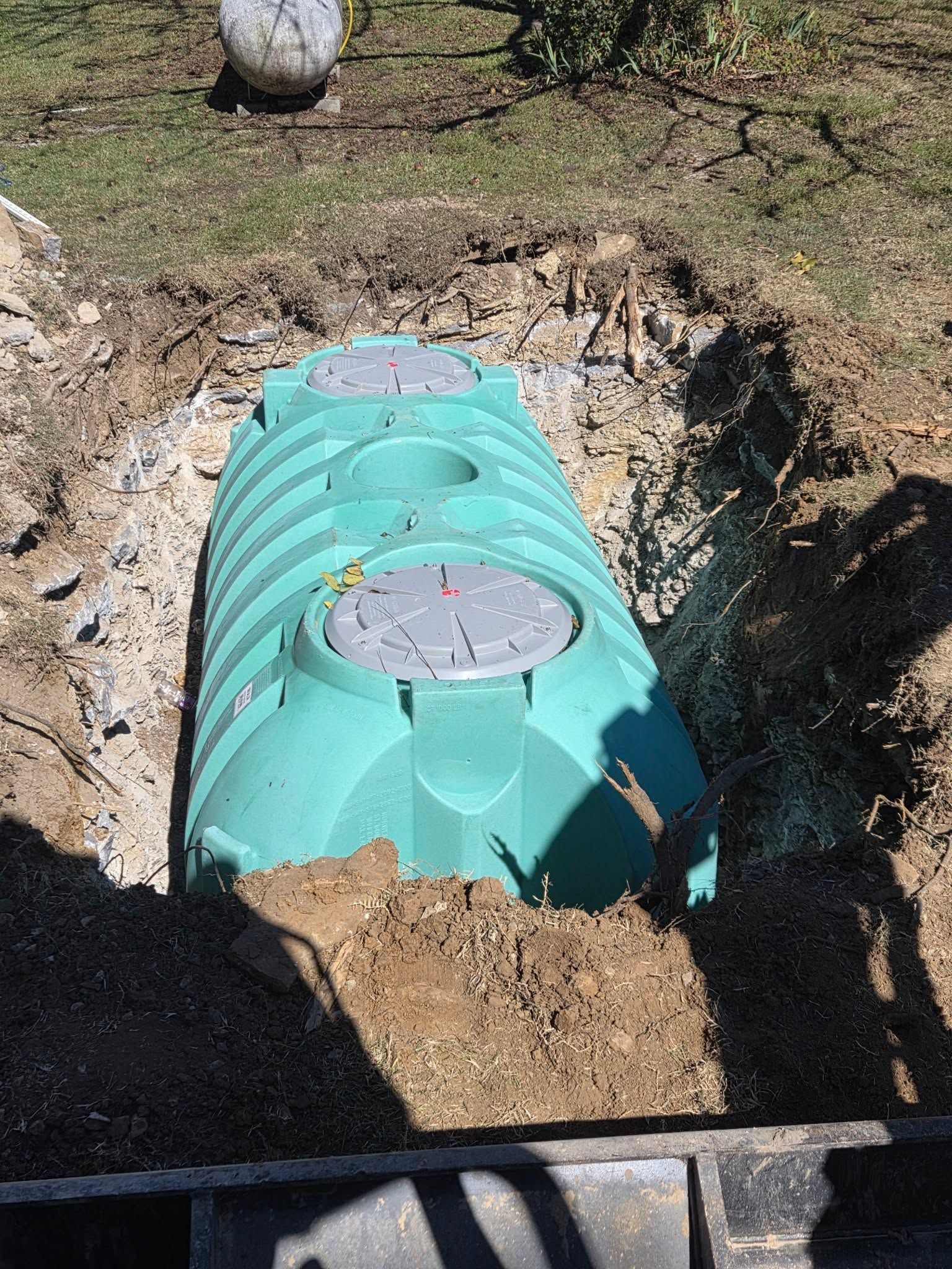 Turquoise underground tank partially buried in dirt, with a round gray access cover on top.