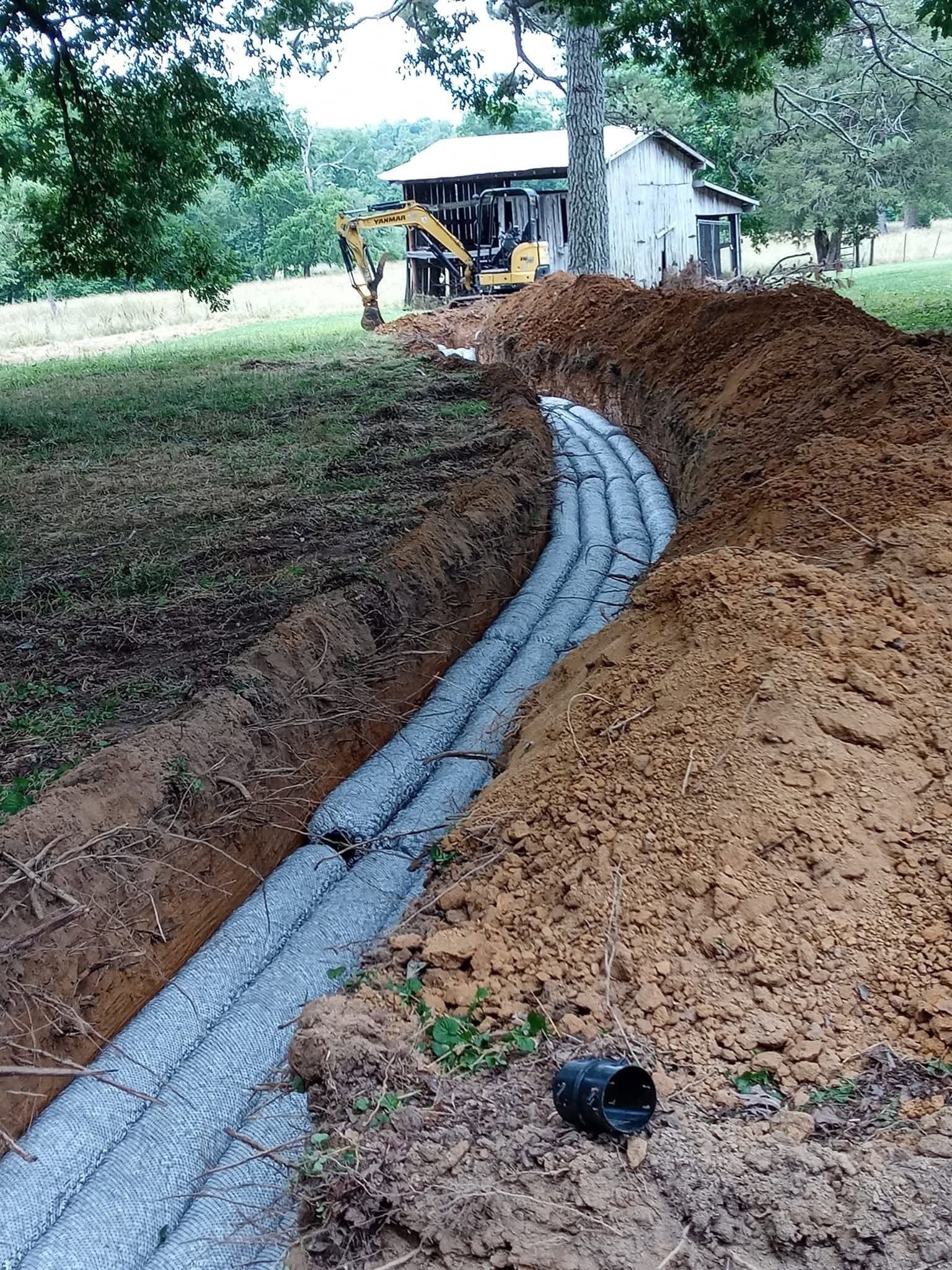 Excavator laying a gray drainage pipe in a dirt trench beside a mound of soil and trees