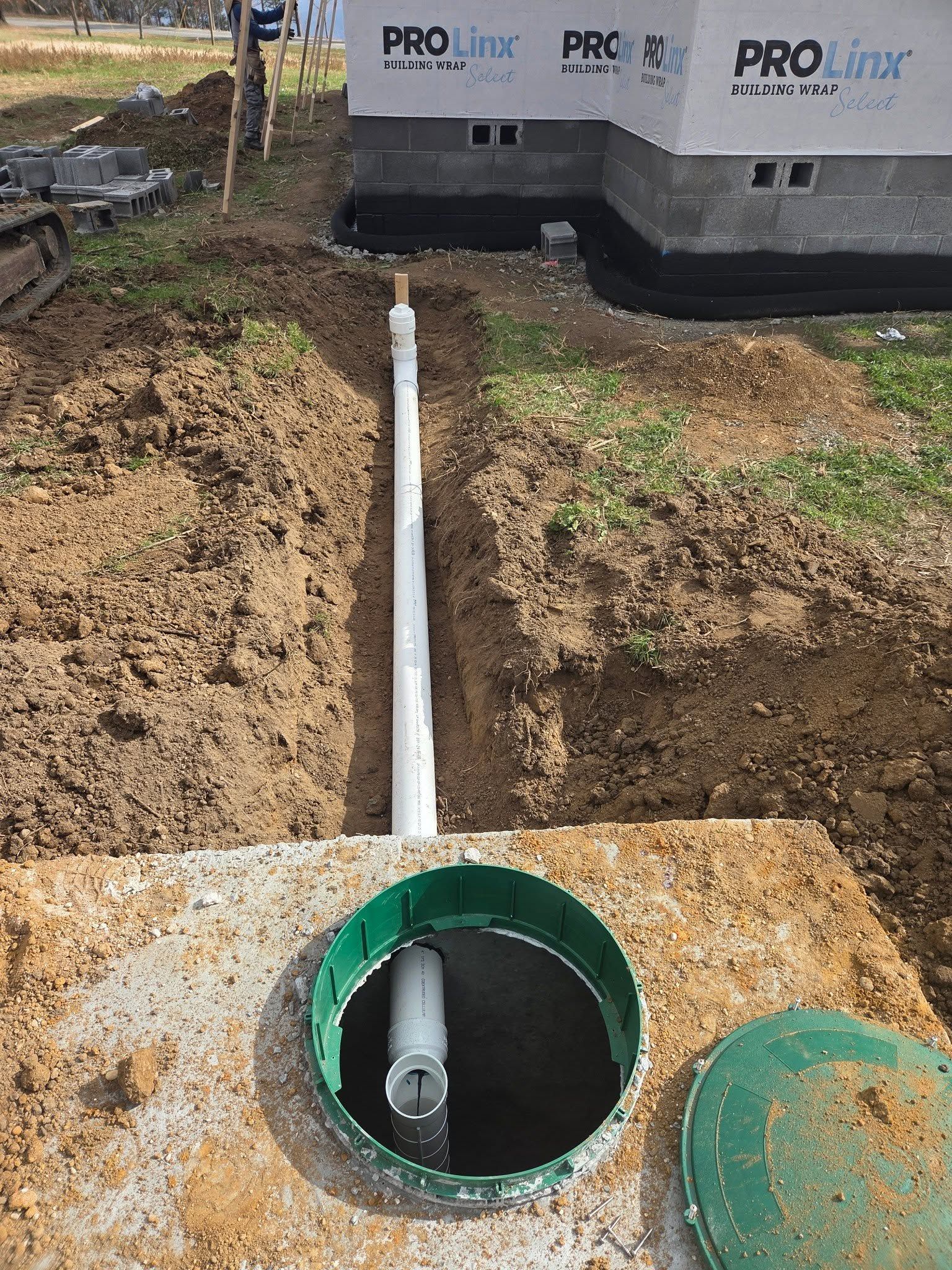 White PVC pipe in a trench leading to a green-capped utility opening near a house foundation.