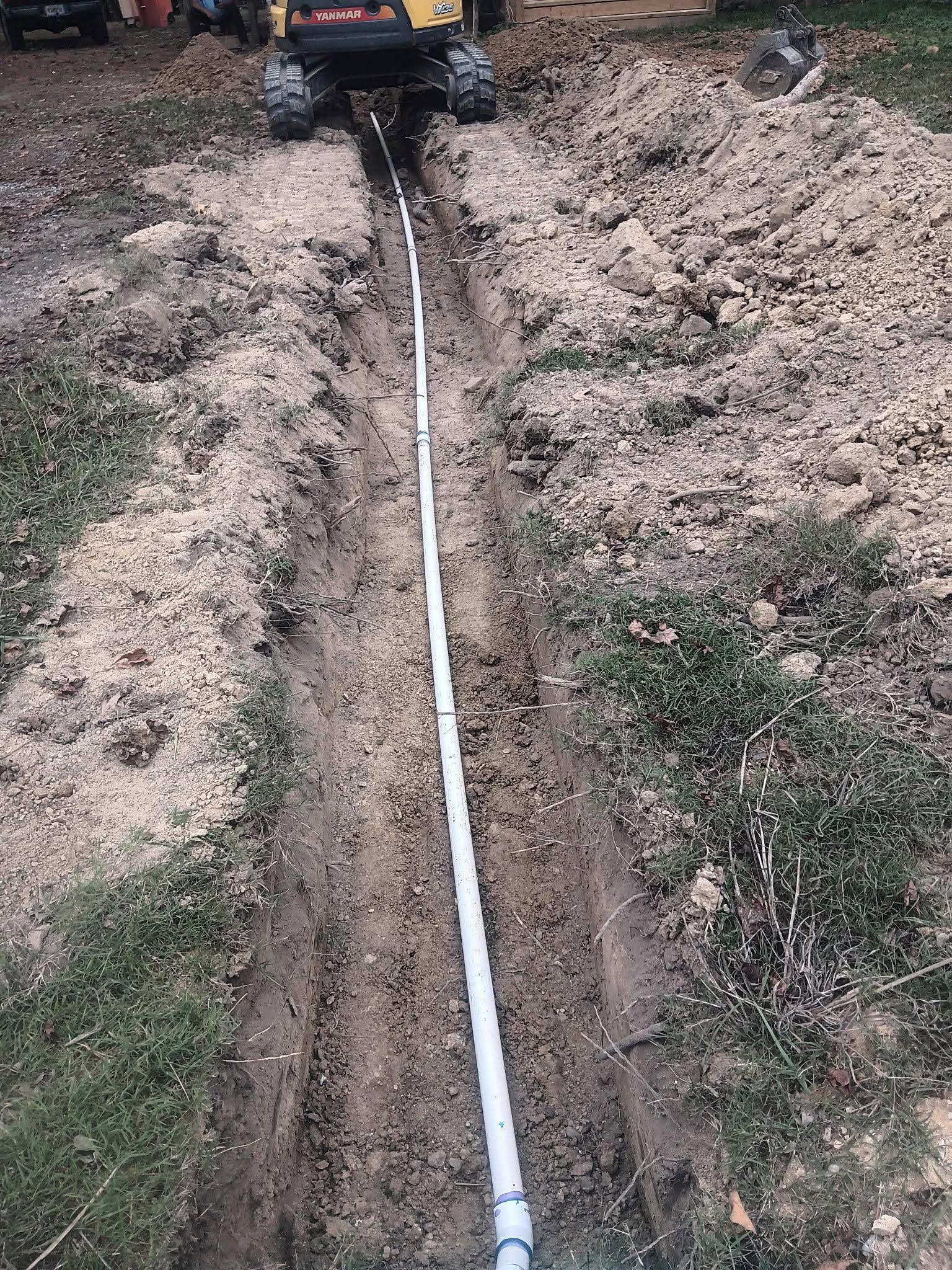 White PVC pipe laid in a dirt trench leading to a vehicle in a muddy yard.