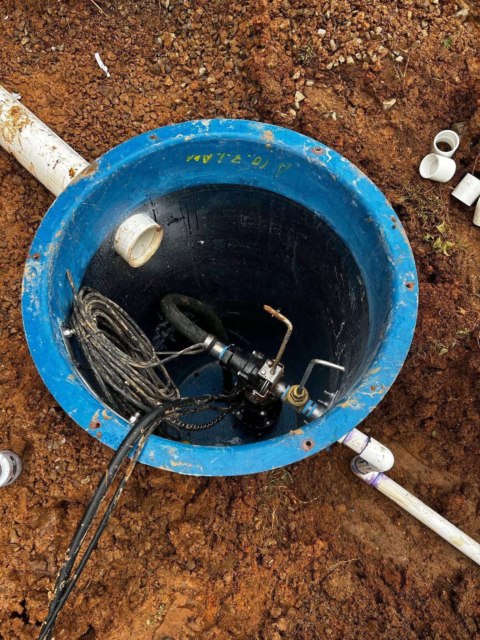 A blue well opening with a black insect trap or device inside, surrounded by dirt and white pipes.