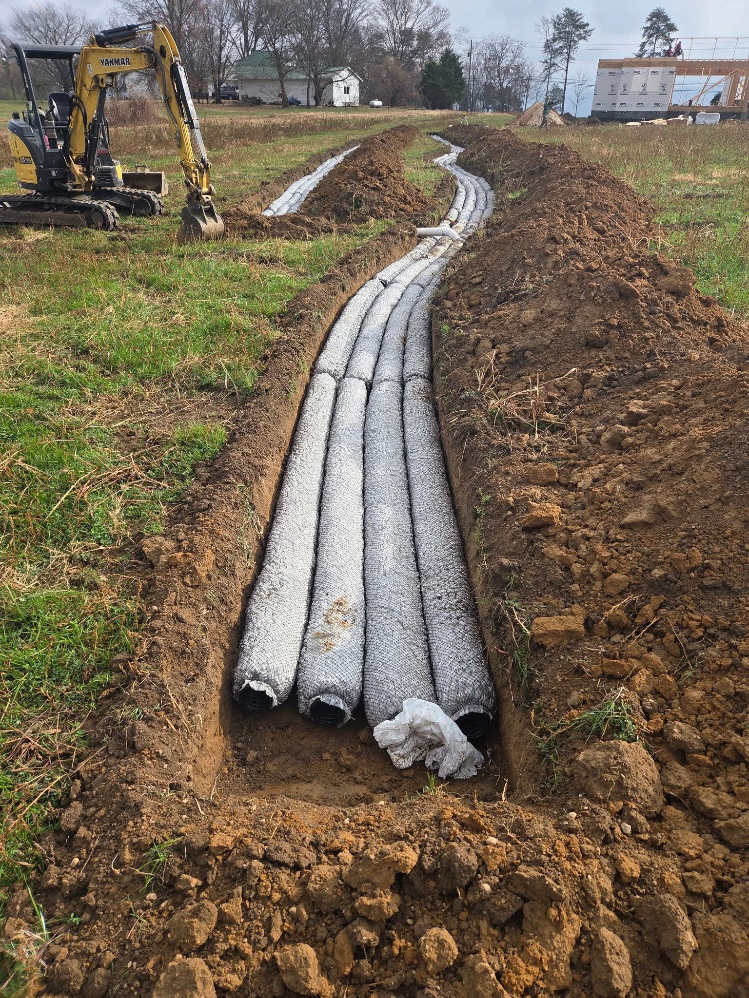 Gray conduit pipes laid in a trench at a muddy construction site, with an excavator nearby.