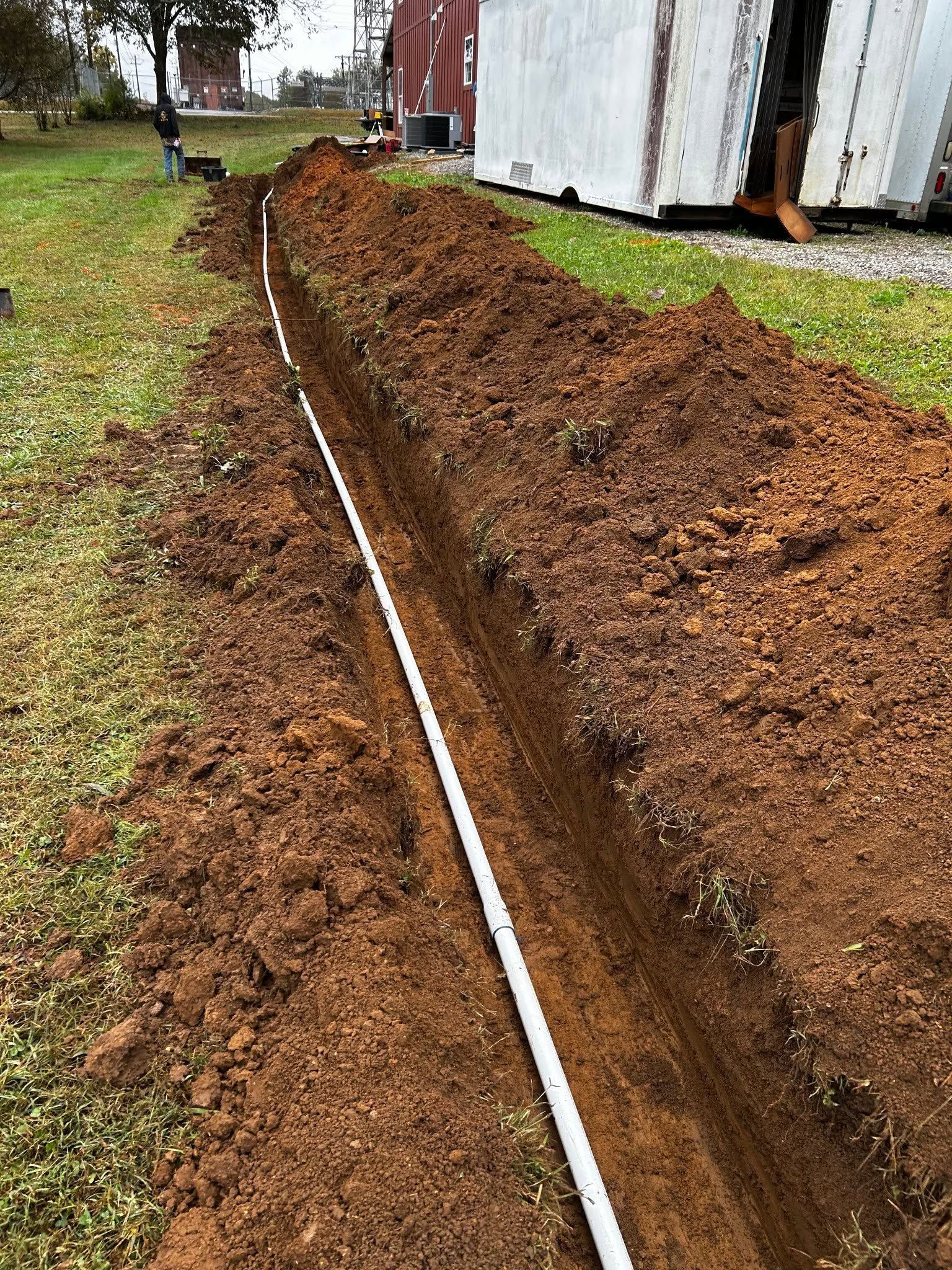Freshly dug trench beside a white building, with a white pipe laid along the dirt