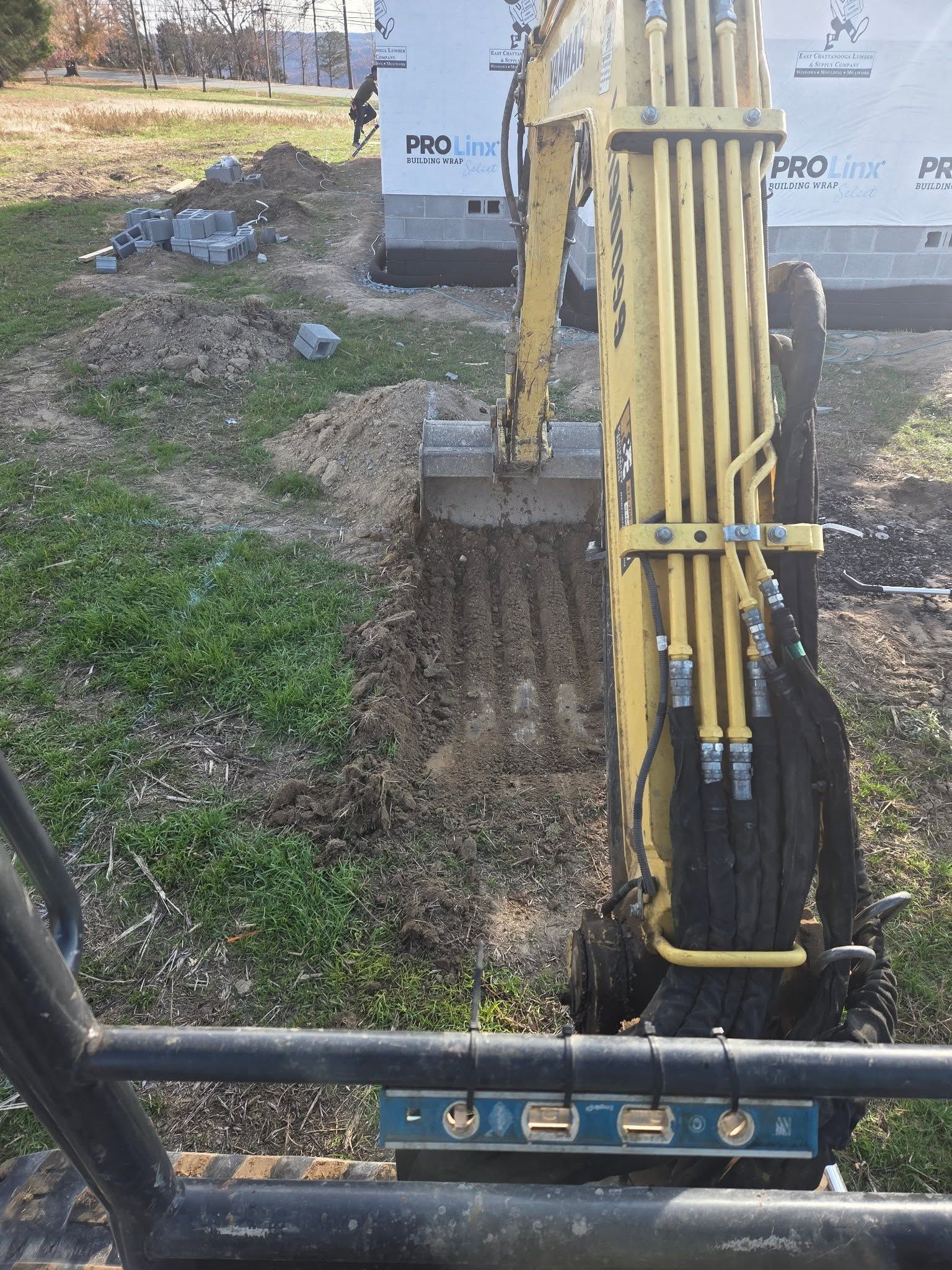 Excavator arm beside a utility box, working in a grassy dirt trench area.
