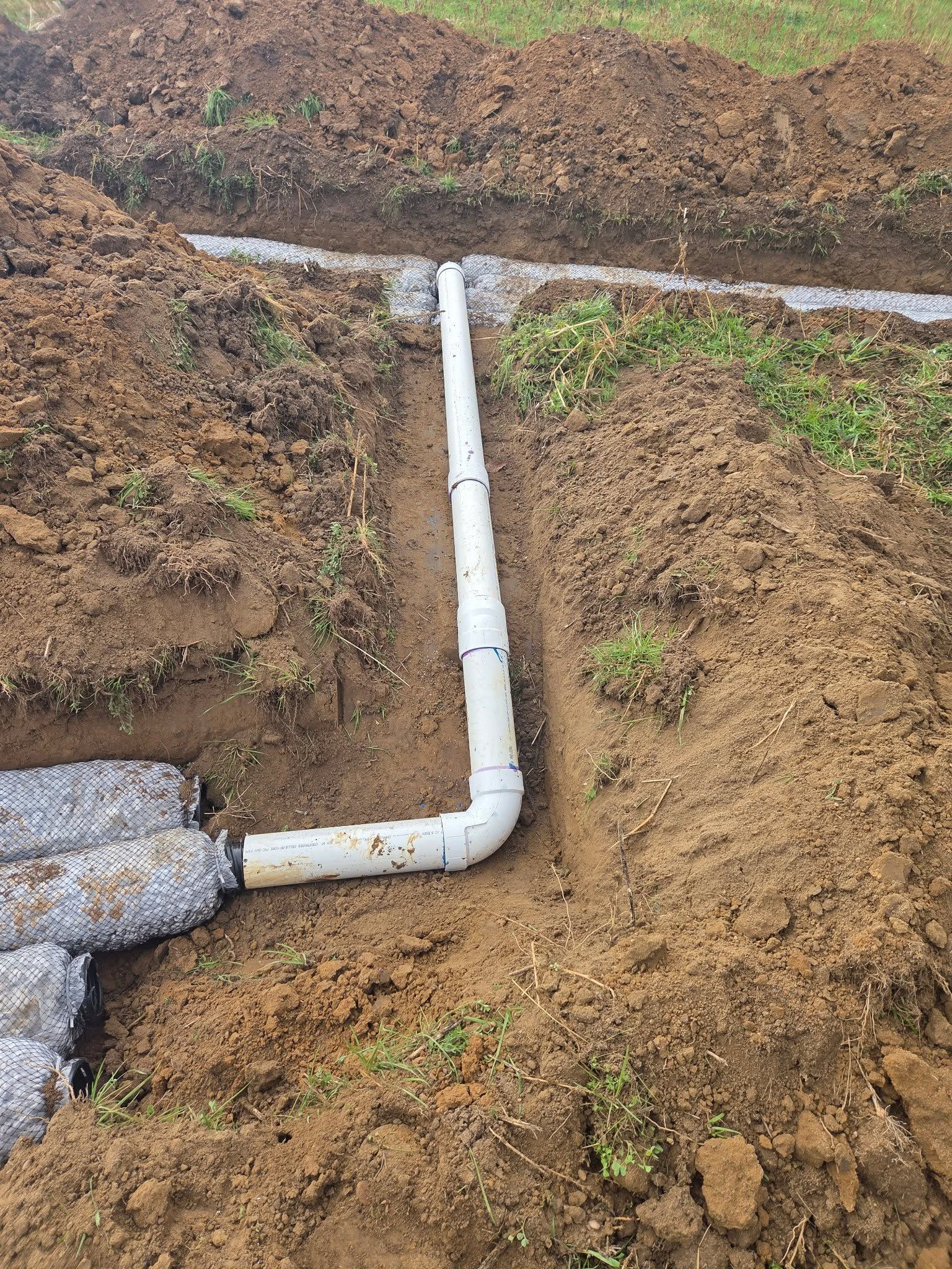 White PVC pipe elbow installed in a dirt trench beside exposed earth.