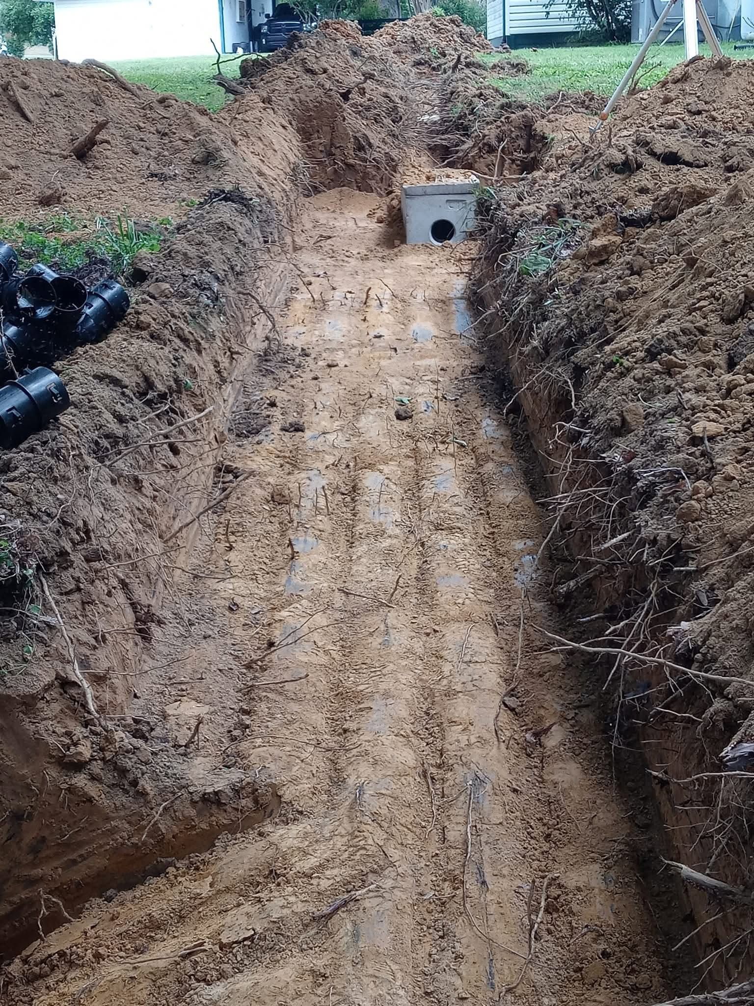 Freshly dug trench lined with wet soil, leading to a small concrete box or drain opening at the far end.