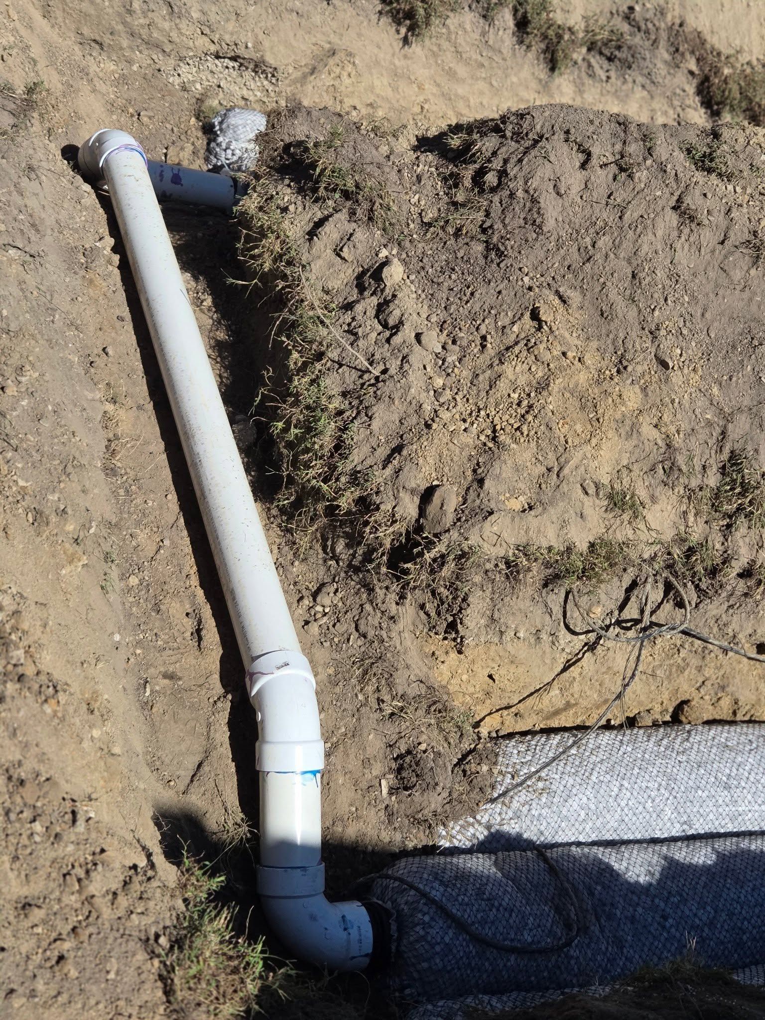 White PVC pipe and black elbow fitting installed in a dirt trench outdoors.