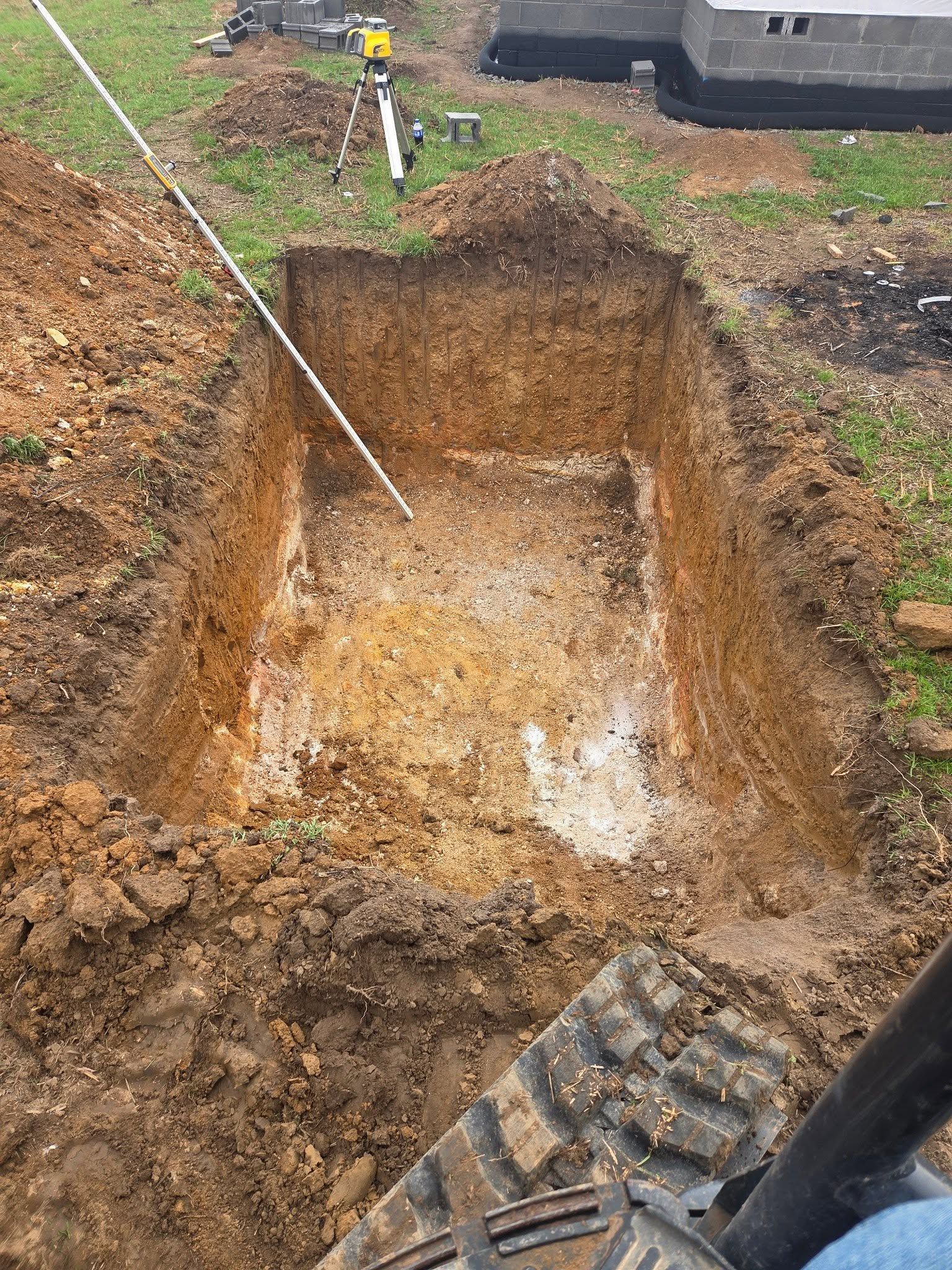 Excavated rectangular hole in muddy ground at a construction site, with surveying equipment nearby.