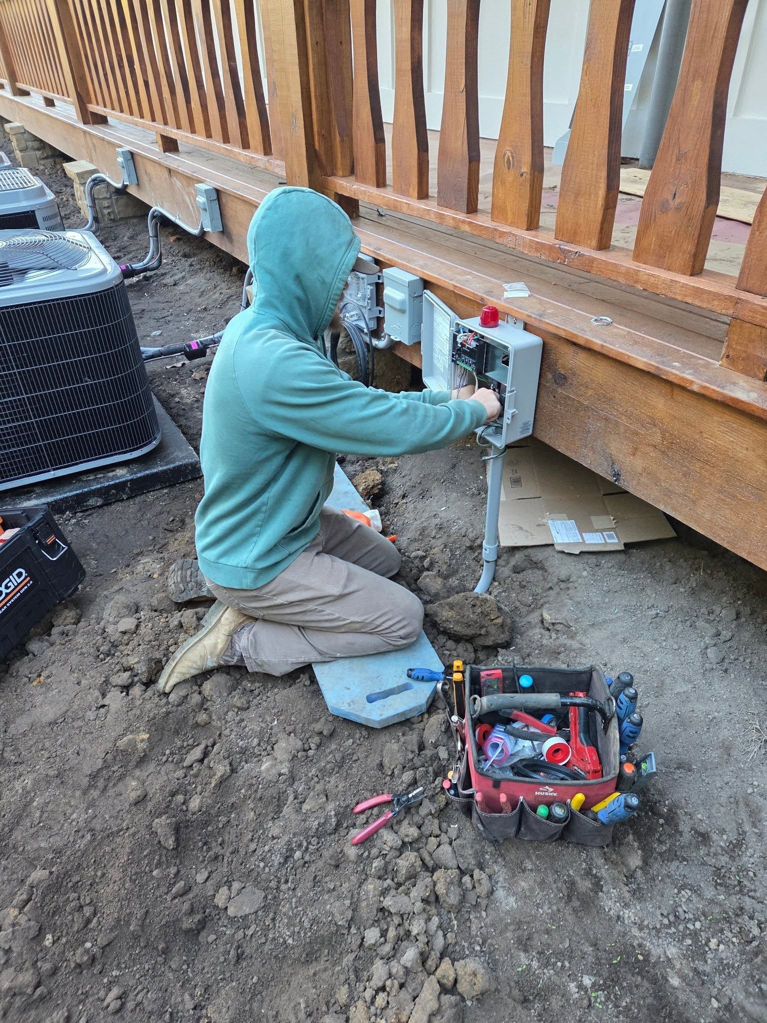 Worker kneeling under a wooden deck, using a power tool on an outdoor electrical box beside tools on muddy ground