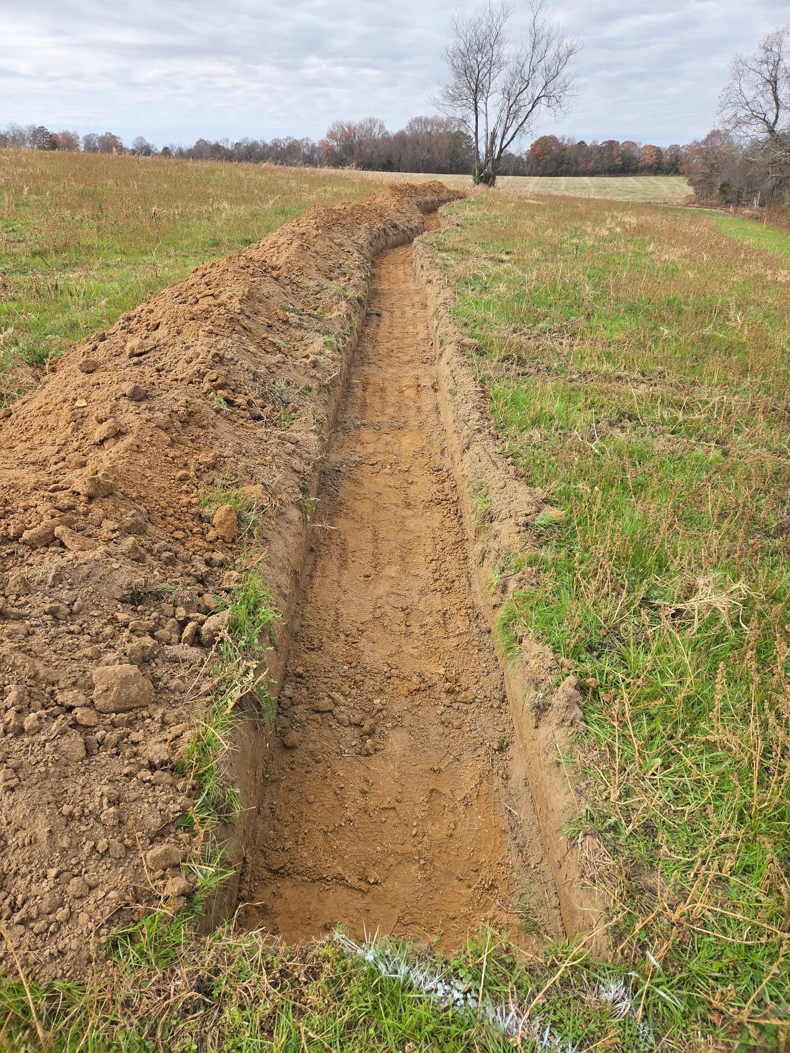 Narrow dirt trench cut through a grassy field under a cloudy sky