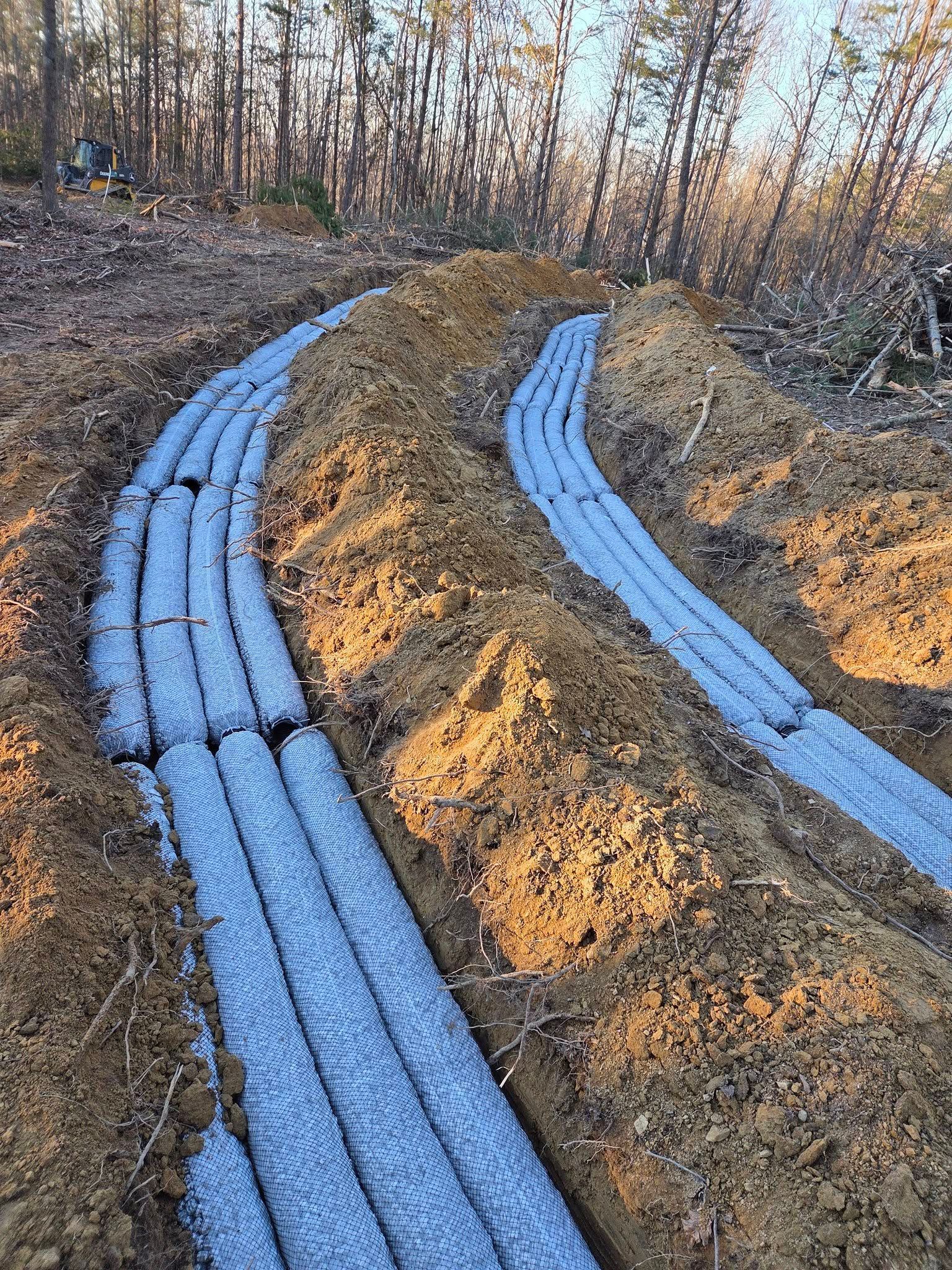 Blue drainage pipes laid in a dirt trench beside a large earth mound in a wooded area