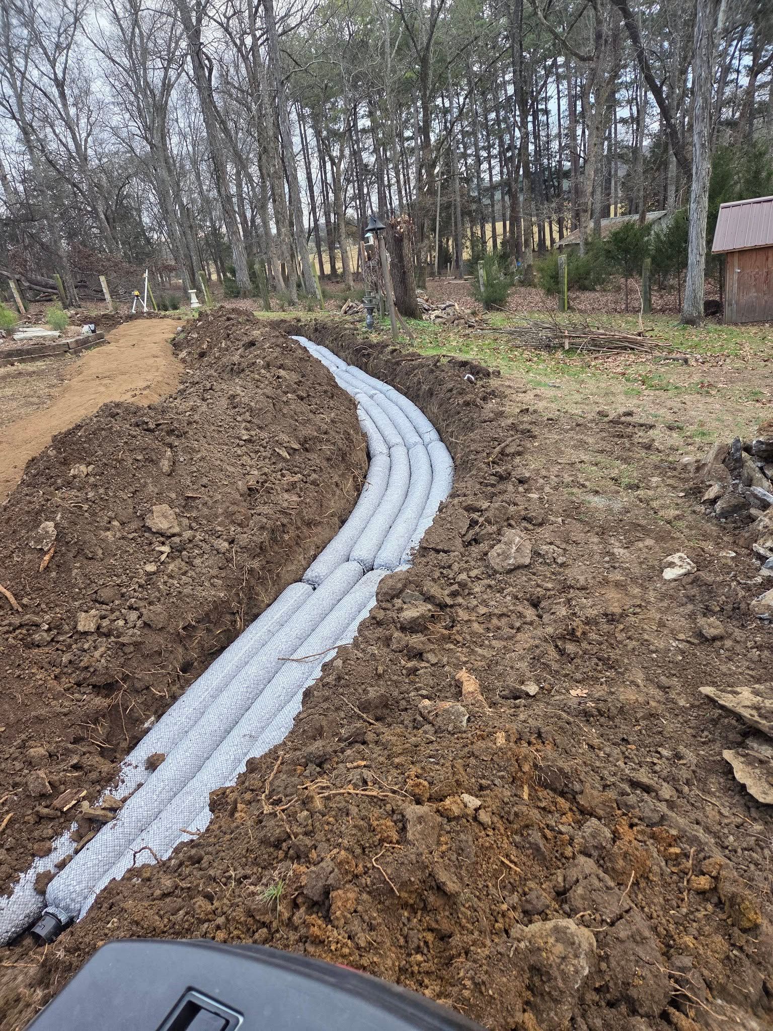 Curved gray drainage pipes laid in a muddy trench beside a dirt path and wooded area
