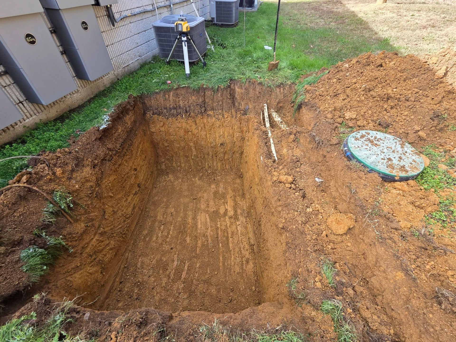 Freshly dug rectangular backyard excavation beside a house, with soil pile and drainage materials nearby