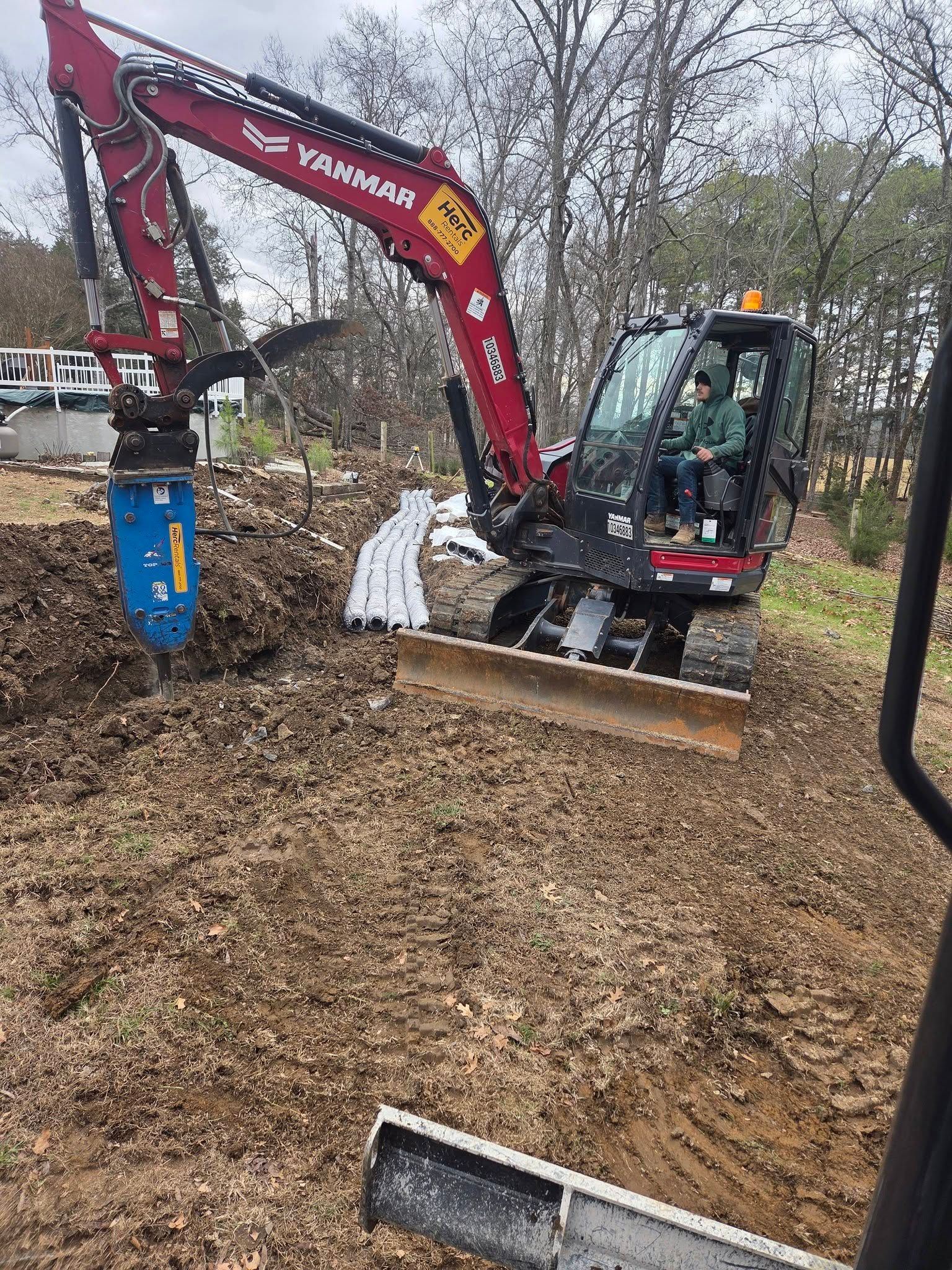 Red excavator clearing leaves in a wooded yard, with a blue auger attachment on the arm.