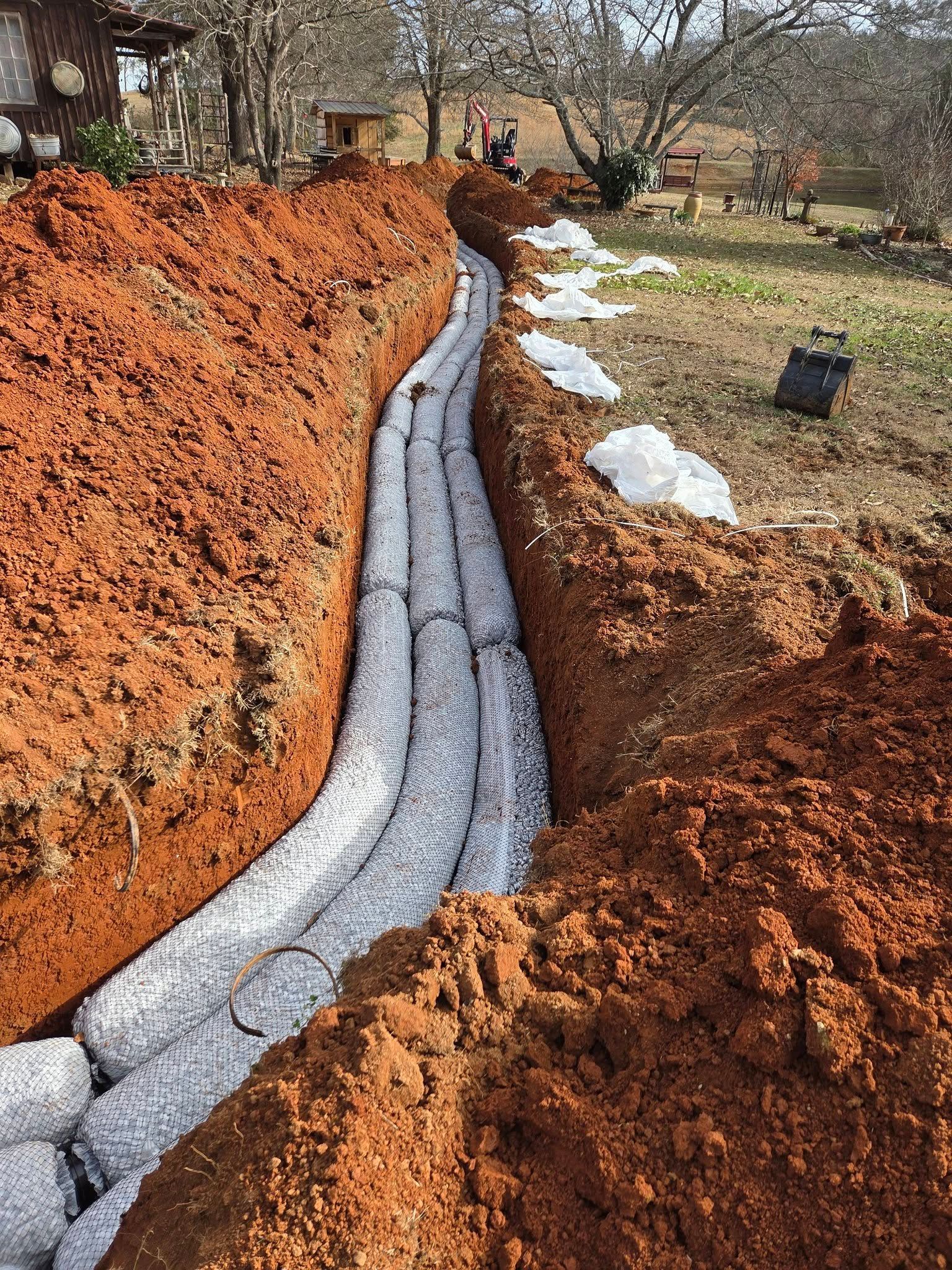 A narrow trench lined with gray drainage pipe runs through red clay in a yard with snow patches.