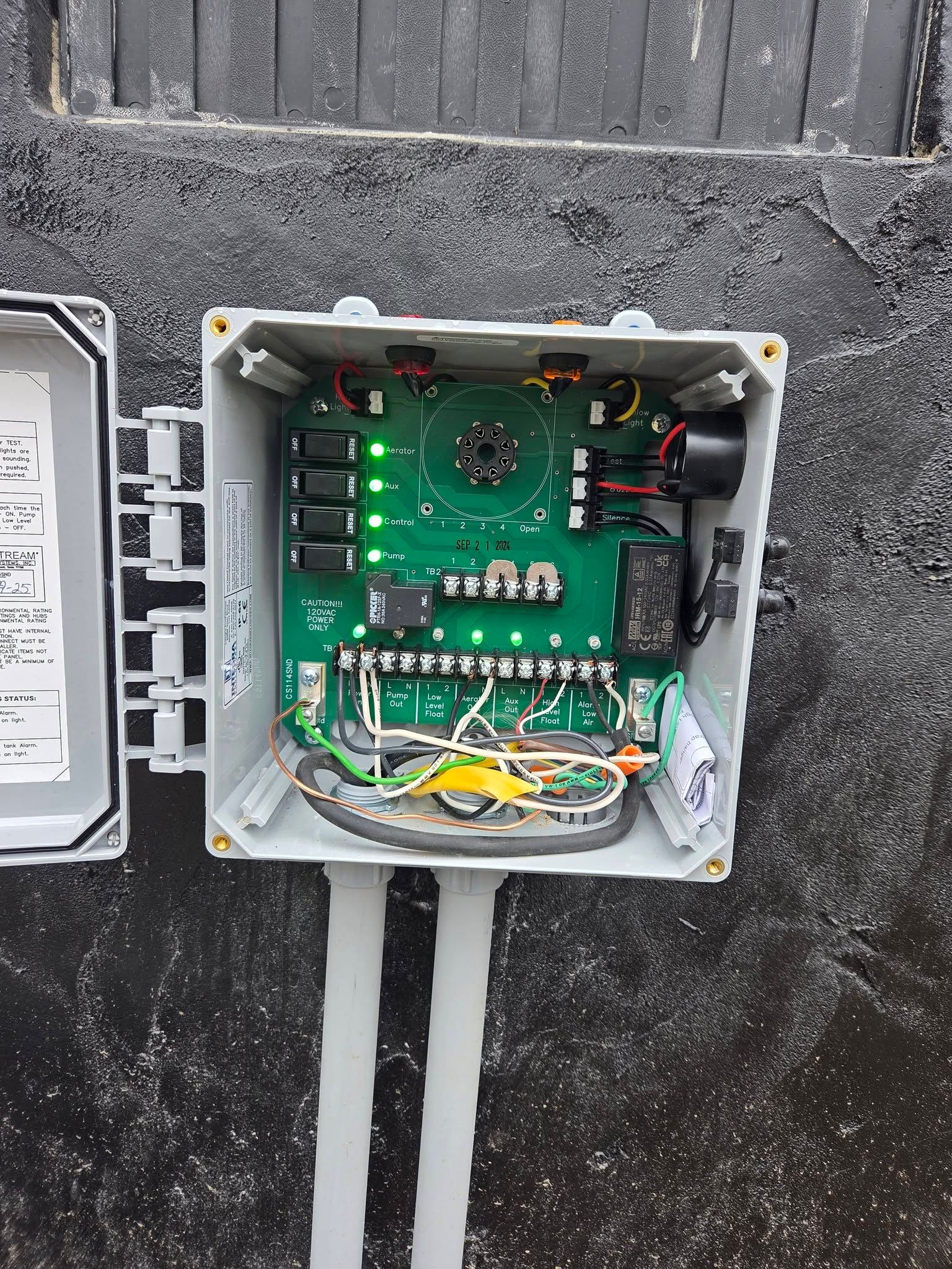 Open electrical junction box with exposed green circuit board and wires on a dark wall.