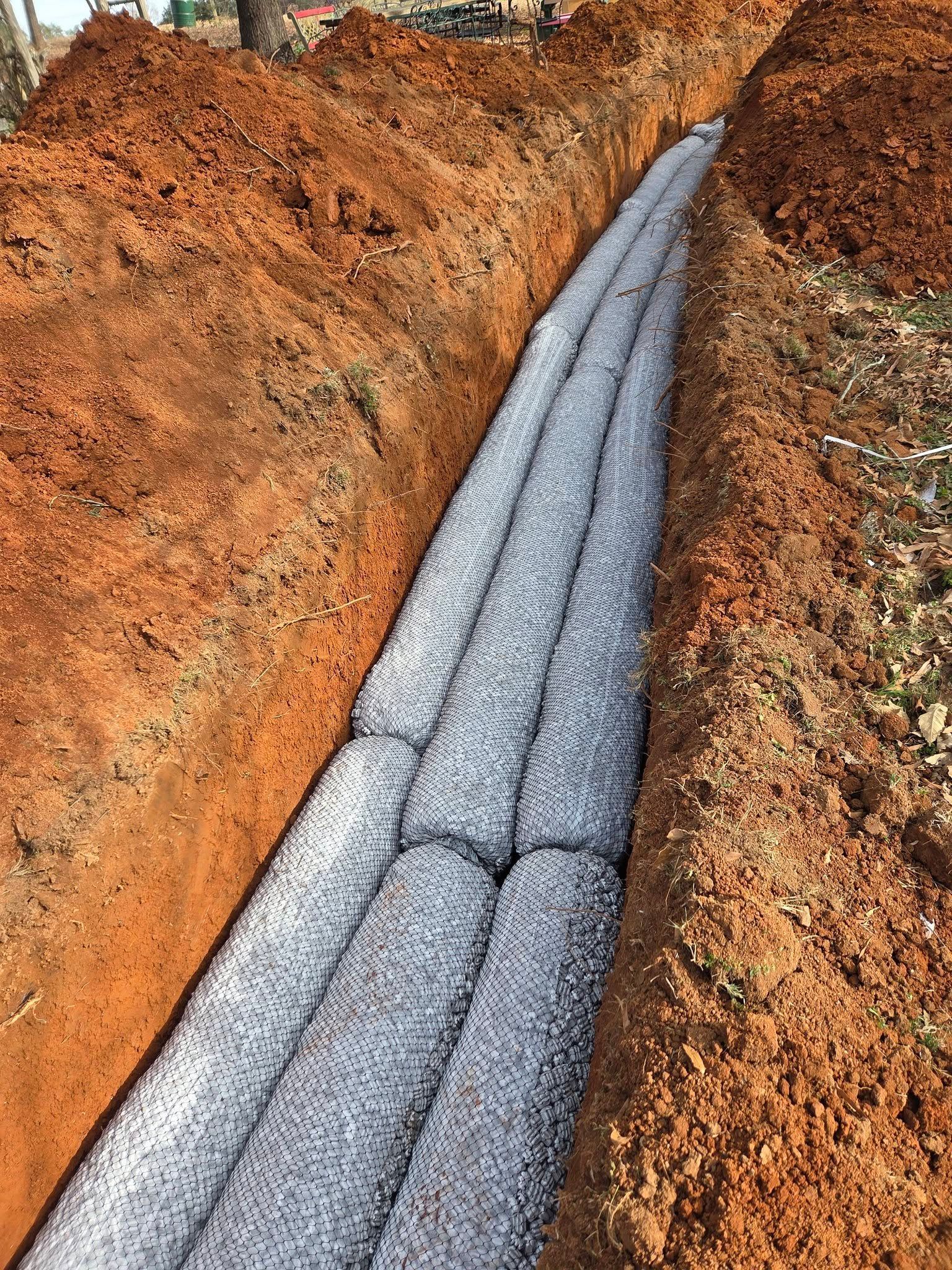 Gray drainage pipe in a reddish dirt trench, with cracked segments running downhill