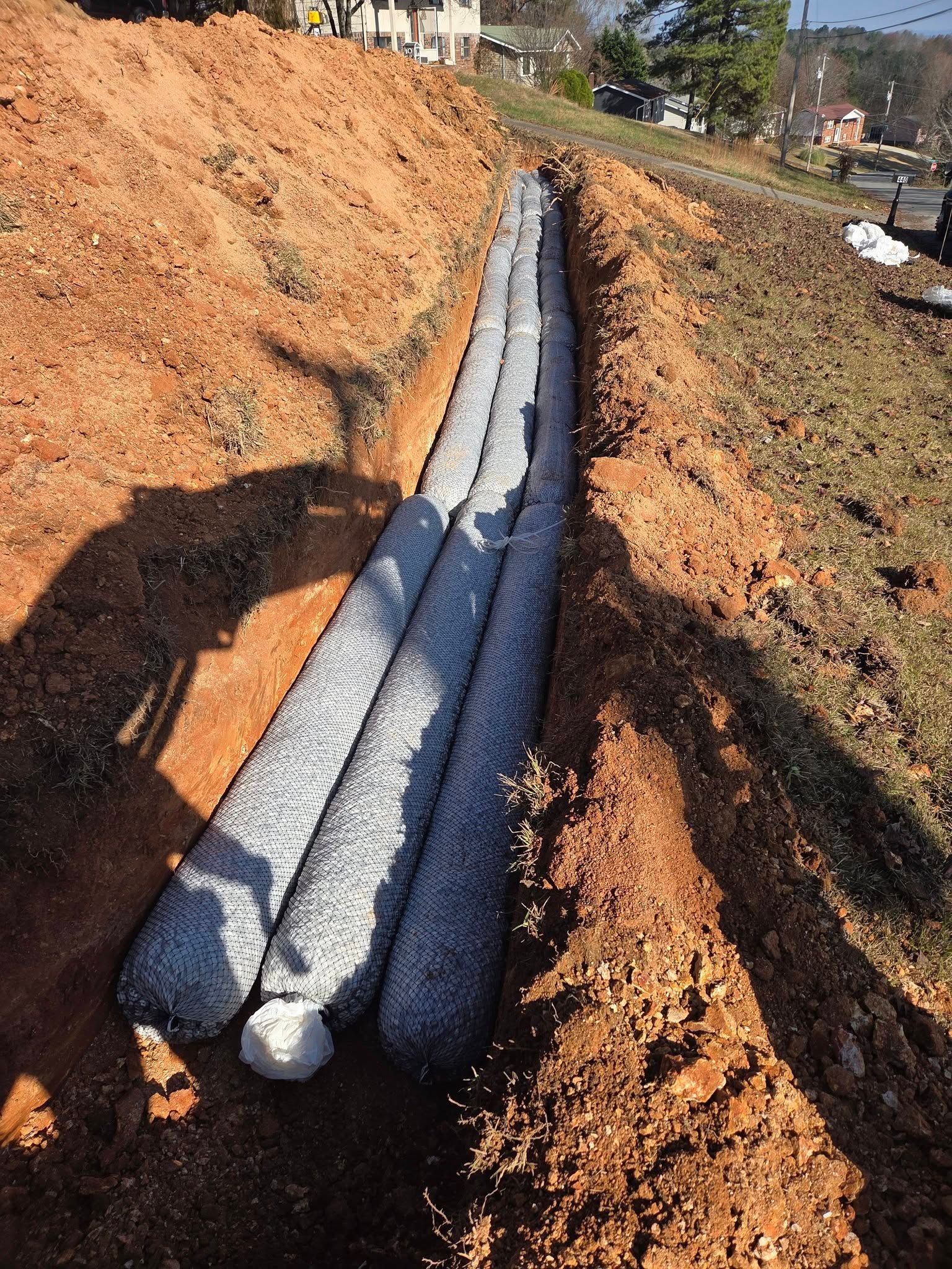 Gray drainage pipes laid in a dirt trench at a construction site, with piles of soil on both sides.