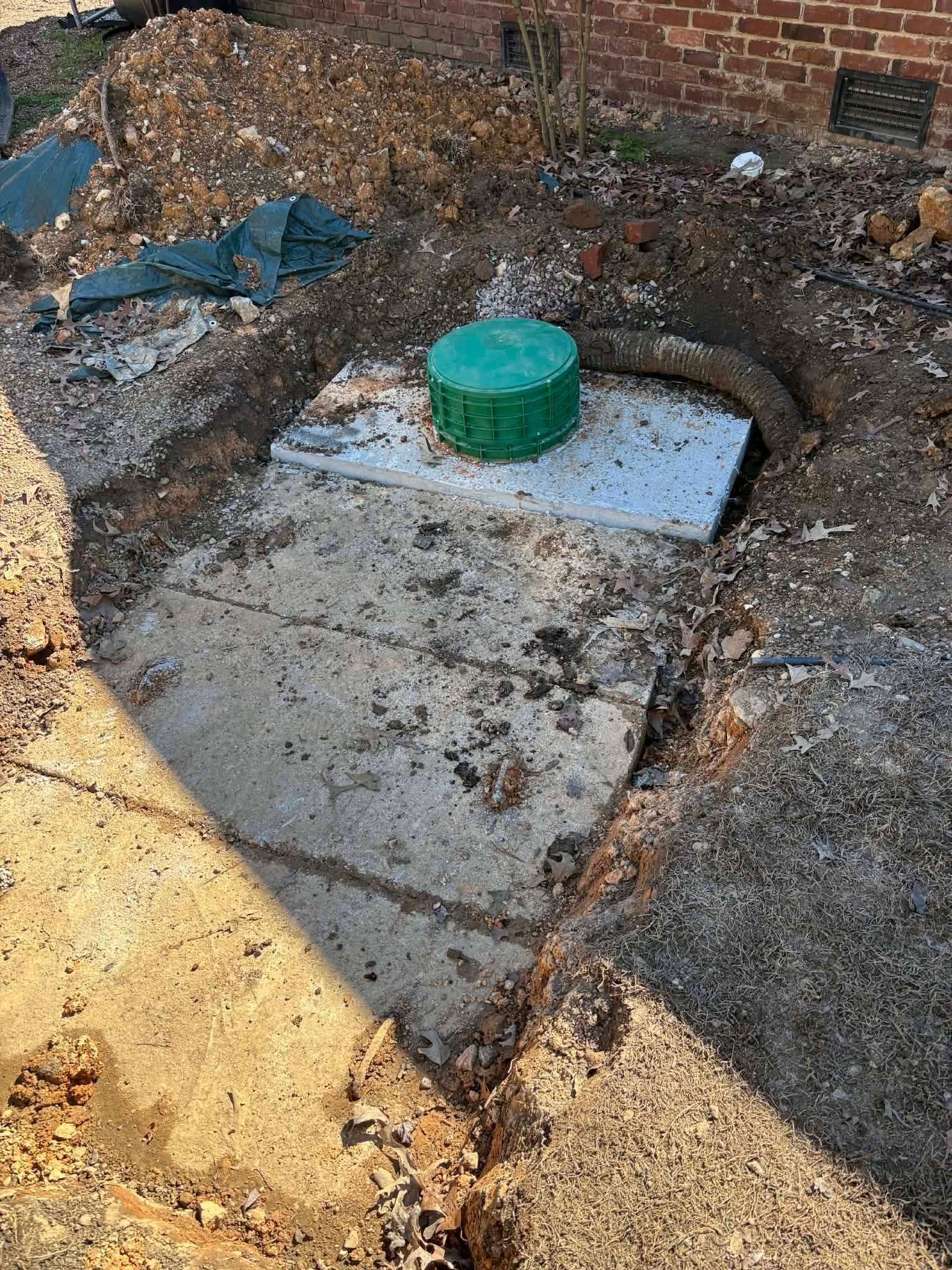 Concrete utility cover with a green cap in a muddy area beside a brick wall