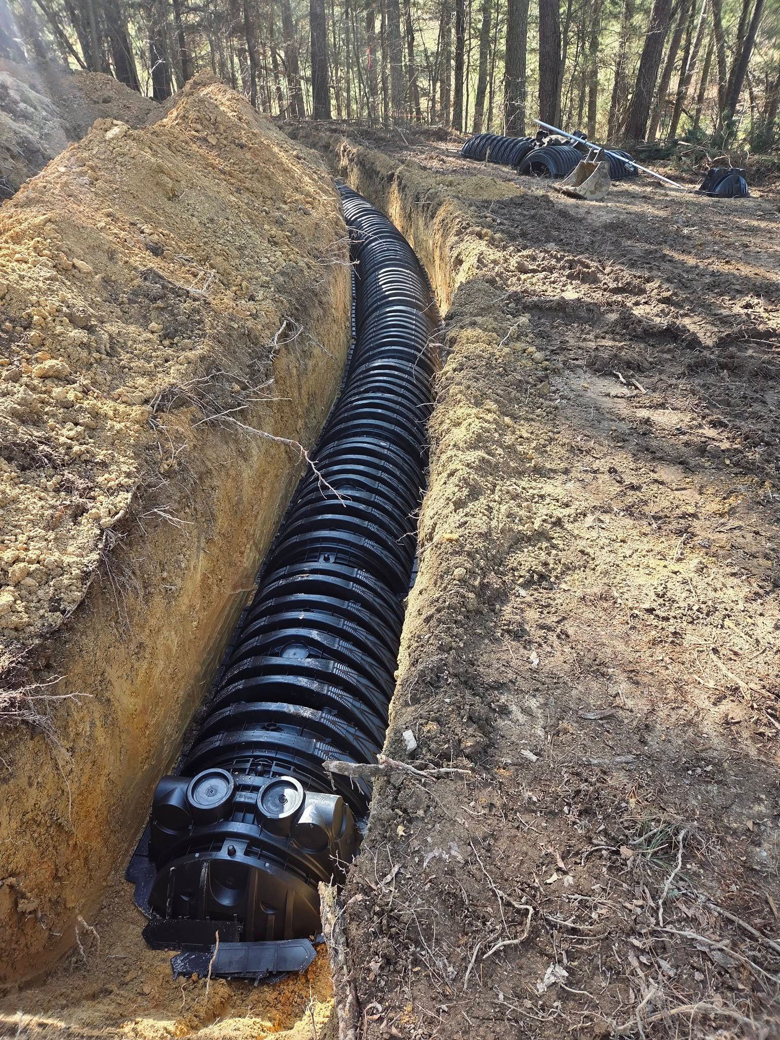 Black corrugated drainage pipe laid in a trench beside a dirt path in a wooded area
