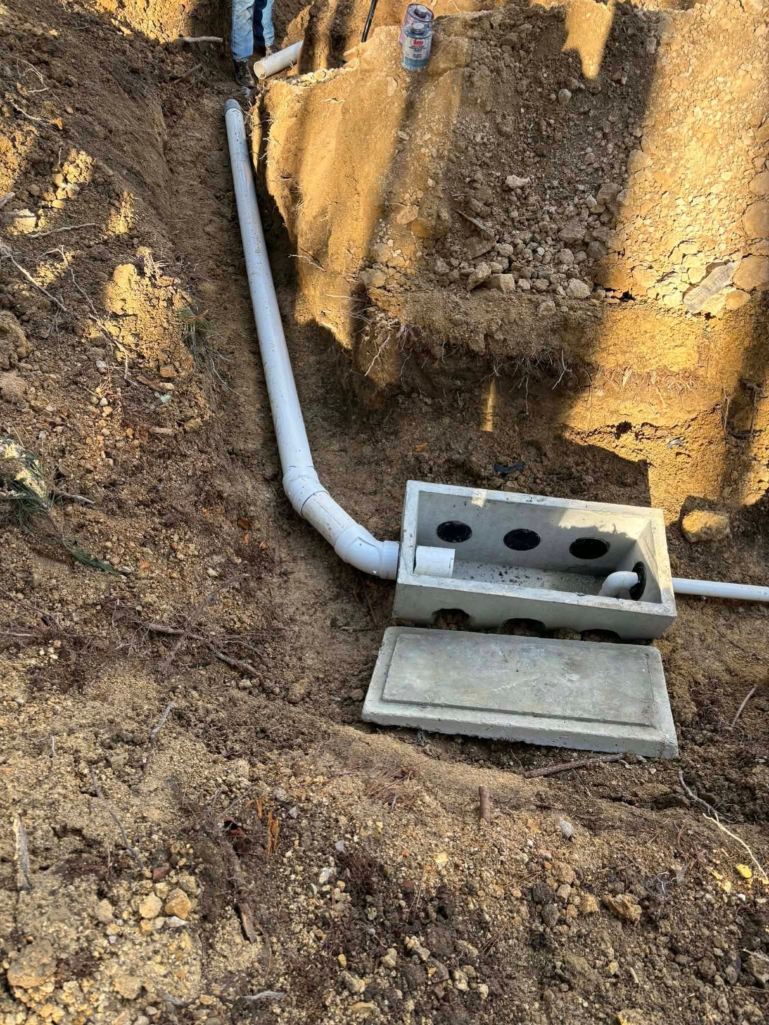 White PVC pipe leading to a gray utility box in a dirt trench beside a concrete wall.
