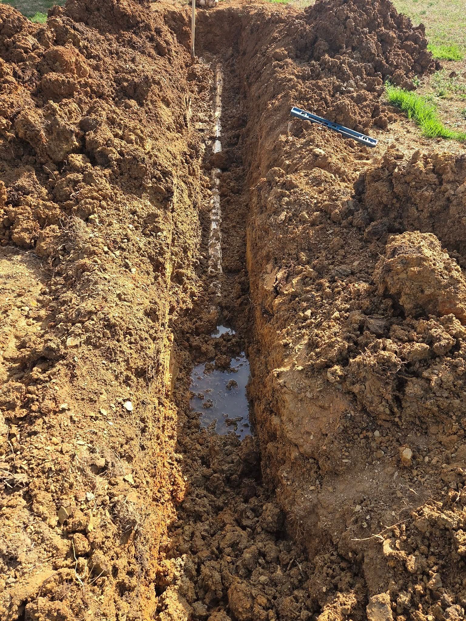Deep muddy trench in soil with pooled water and a blue marker, beside green grass