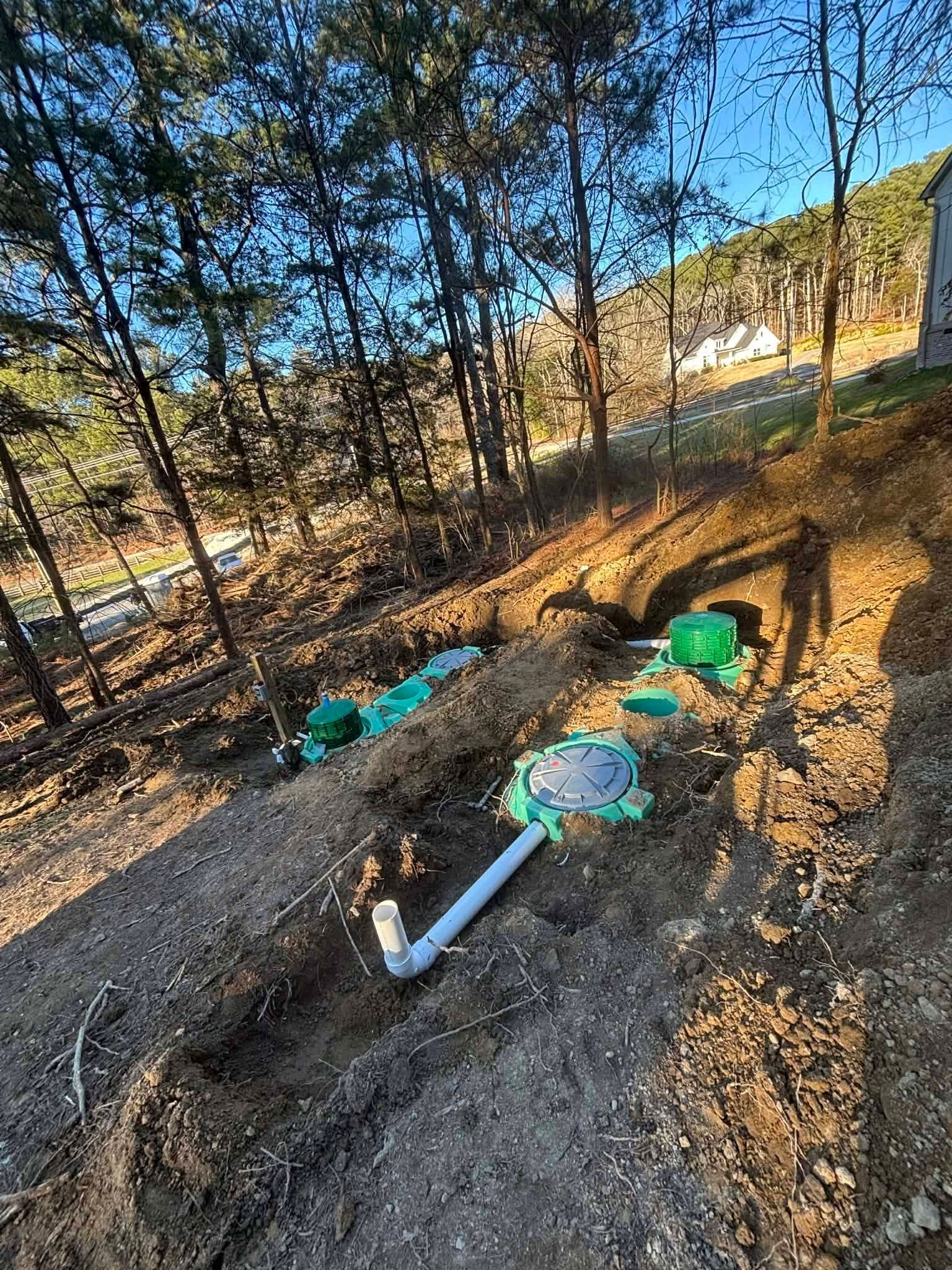 Blue pipe laid in a dirt trench on a wooded hillside, with green fittings and sunlight filtering through trees.