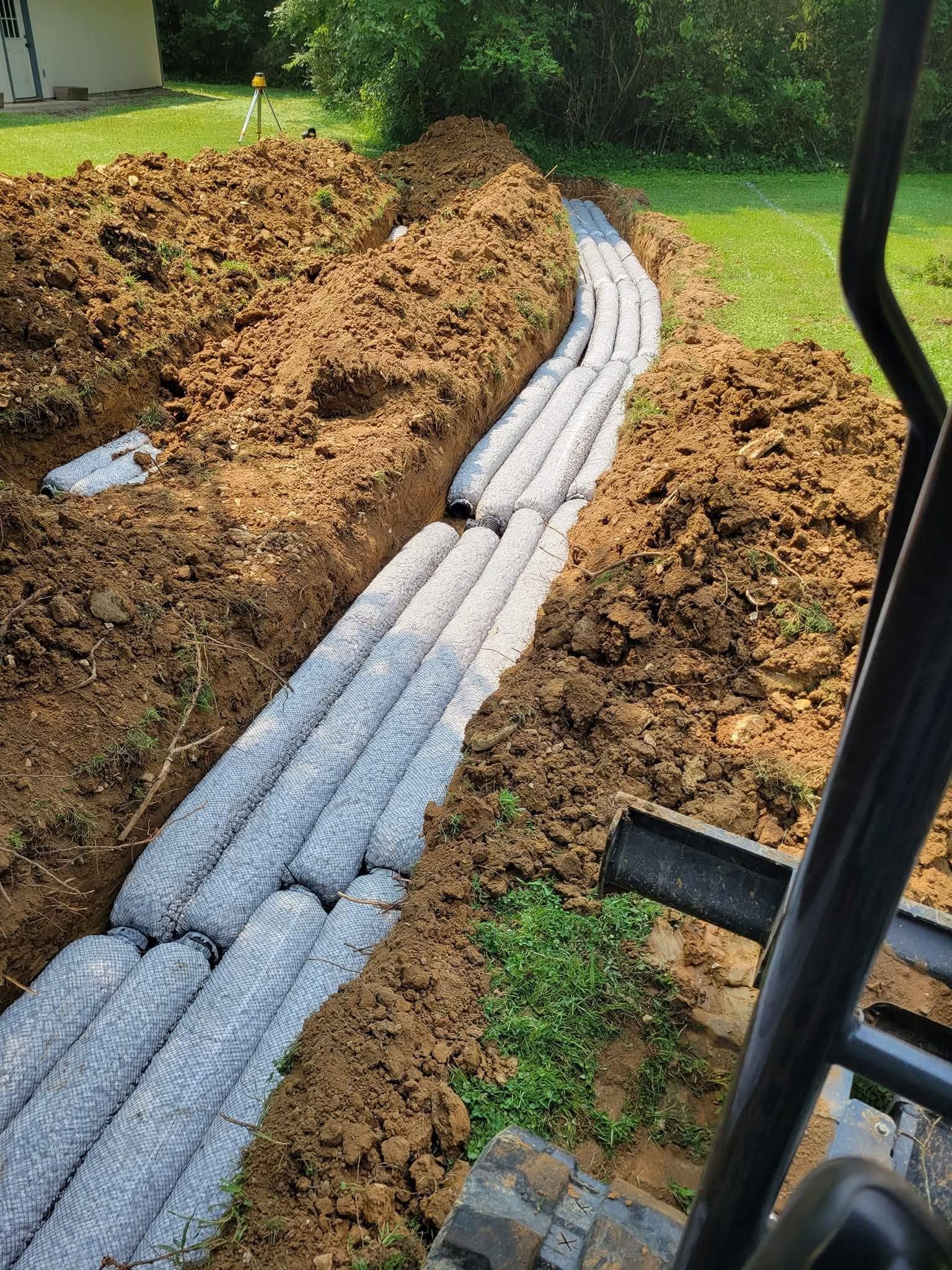 White drainage pipe lined in a trench beside a dirt mound, with a small excavator in the foreground.