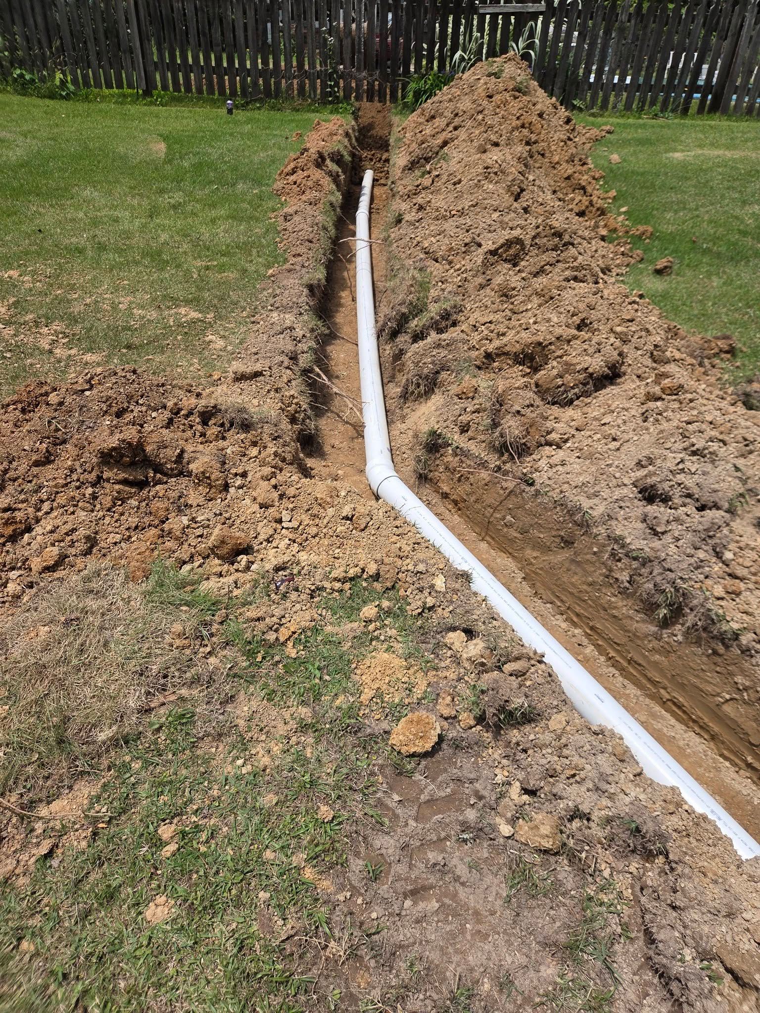 White drainage pipe laid in a trench beside a grassy yard, with piles of excavated soil.