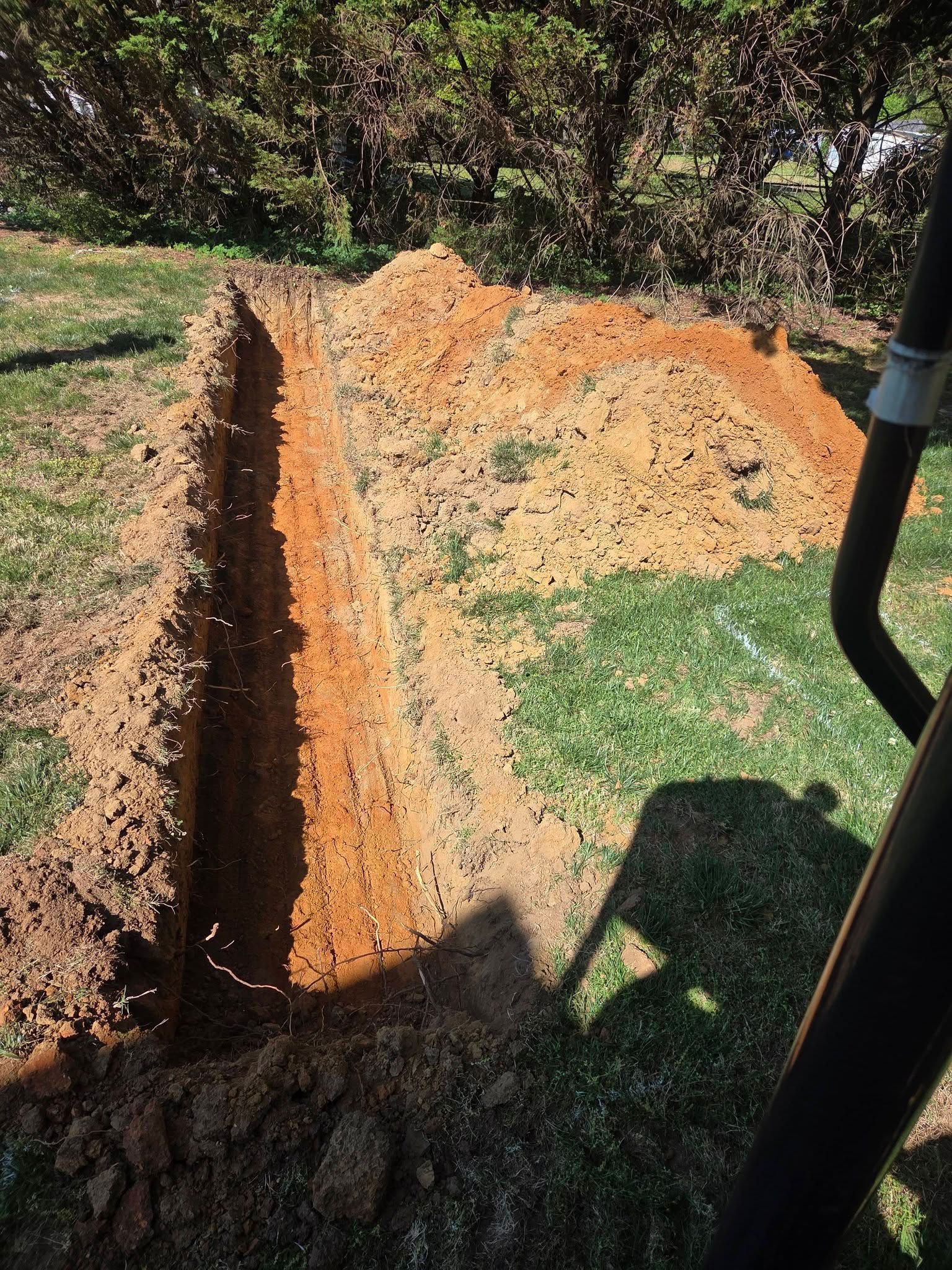 Freshly dug trench in a grassy yard with orange soil and a shovel handle at right