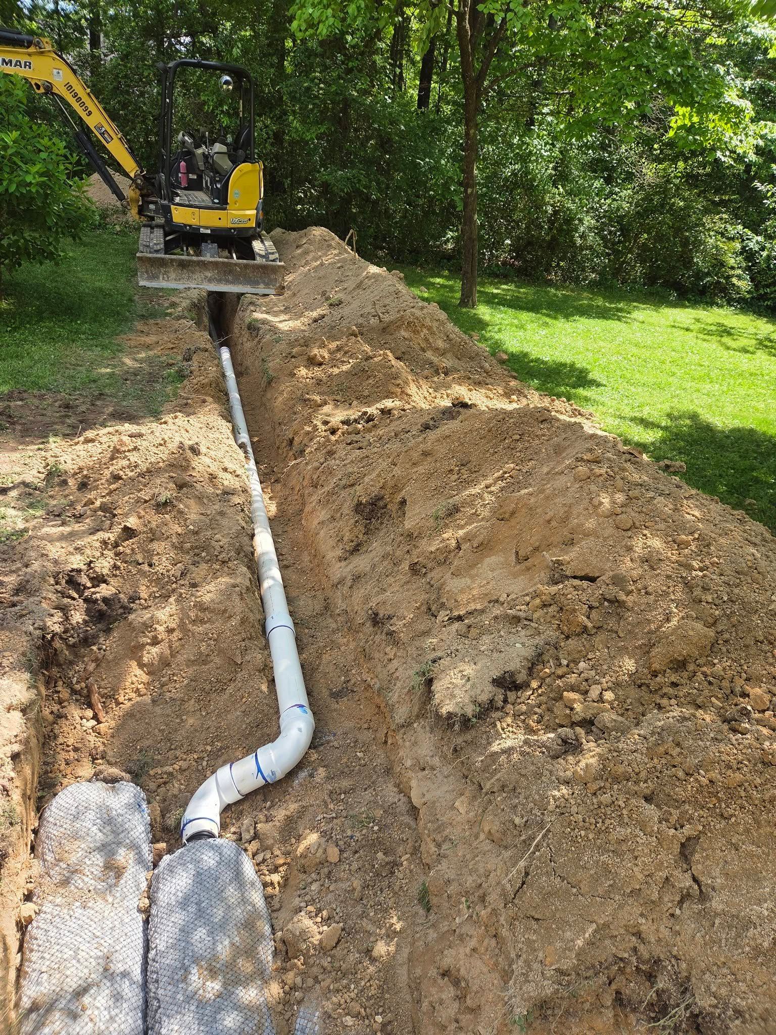 Excavator trenching beside a white pipe laid in a dirt mound along a wooded yard