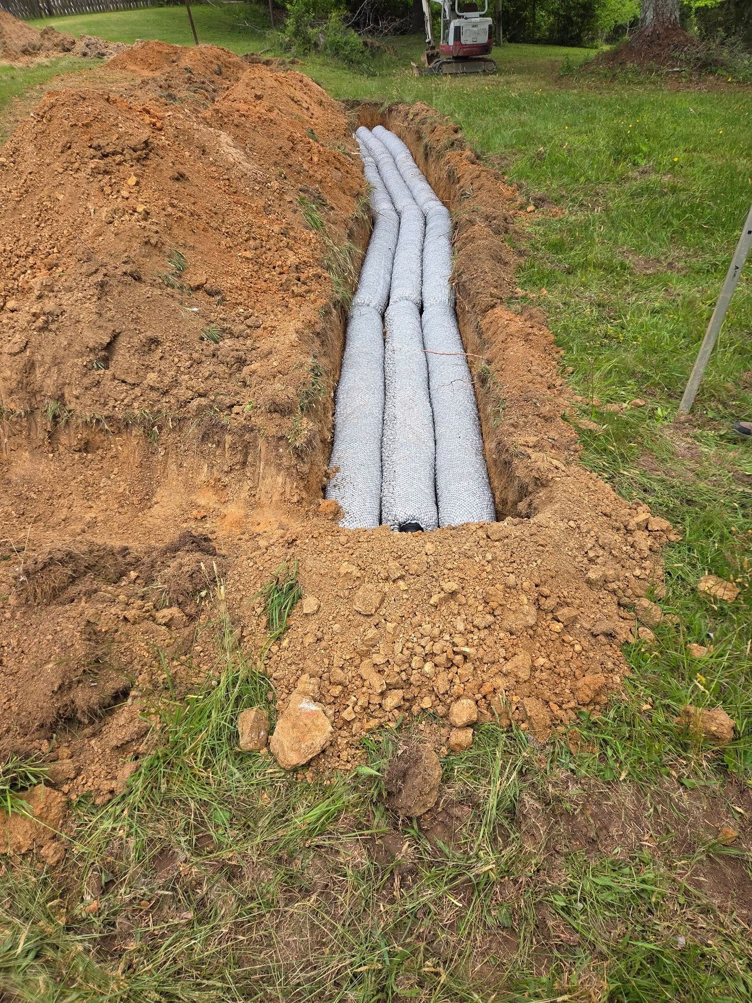 Shredded wood chips piled around a narrow trench lined with gray drainage pipe outdoors.