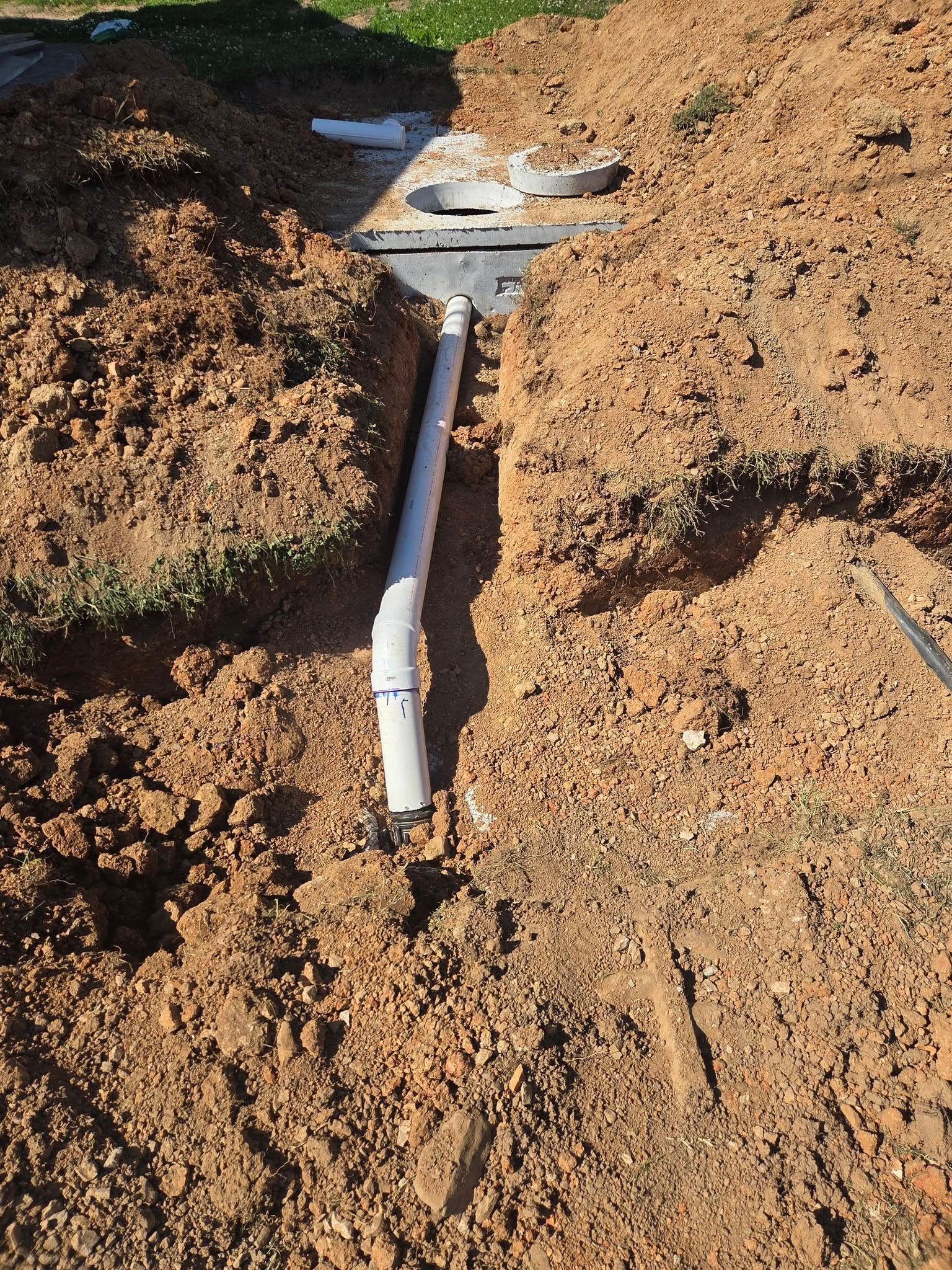 White PVC pipe trench in brown dirt, with exposed plumbing line running through excavated ground.