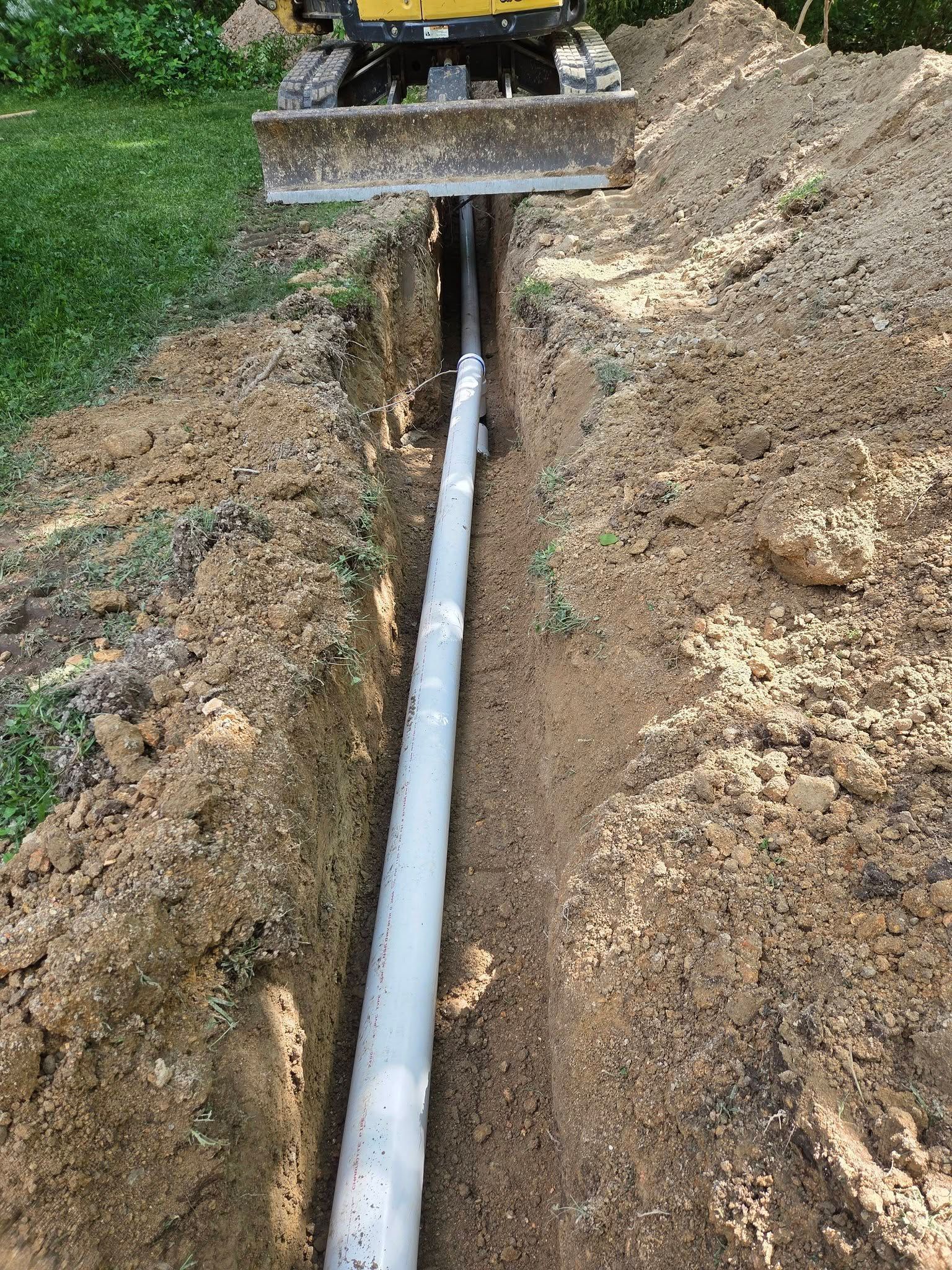 A trench with a white PVC pipe being laid under soil by a construction machine.