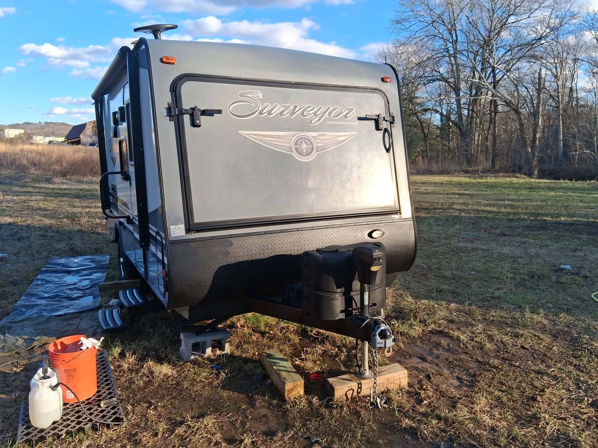 Gray travel trailer parked on grass with a front hitch, under a blue sky near leafless trees