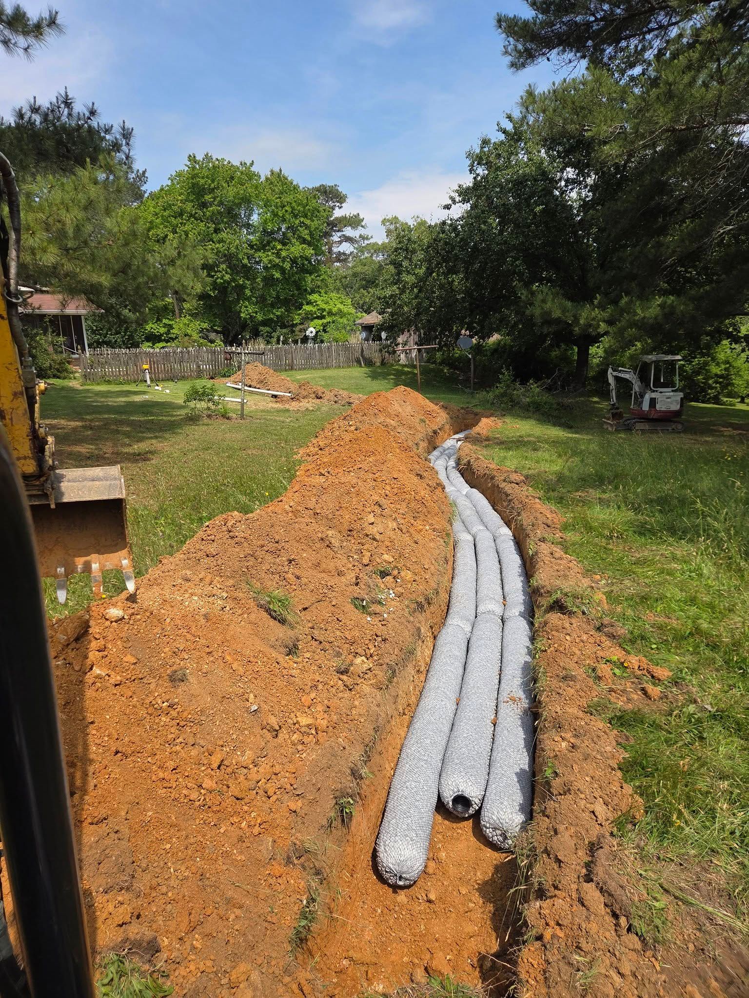 Orange soil mound with two gray pipes laid on a grassy path beside trees and a parked vehicle