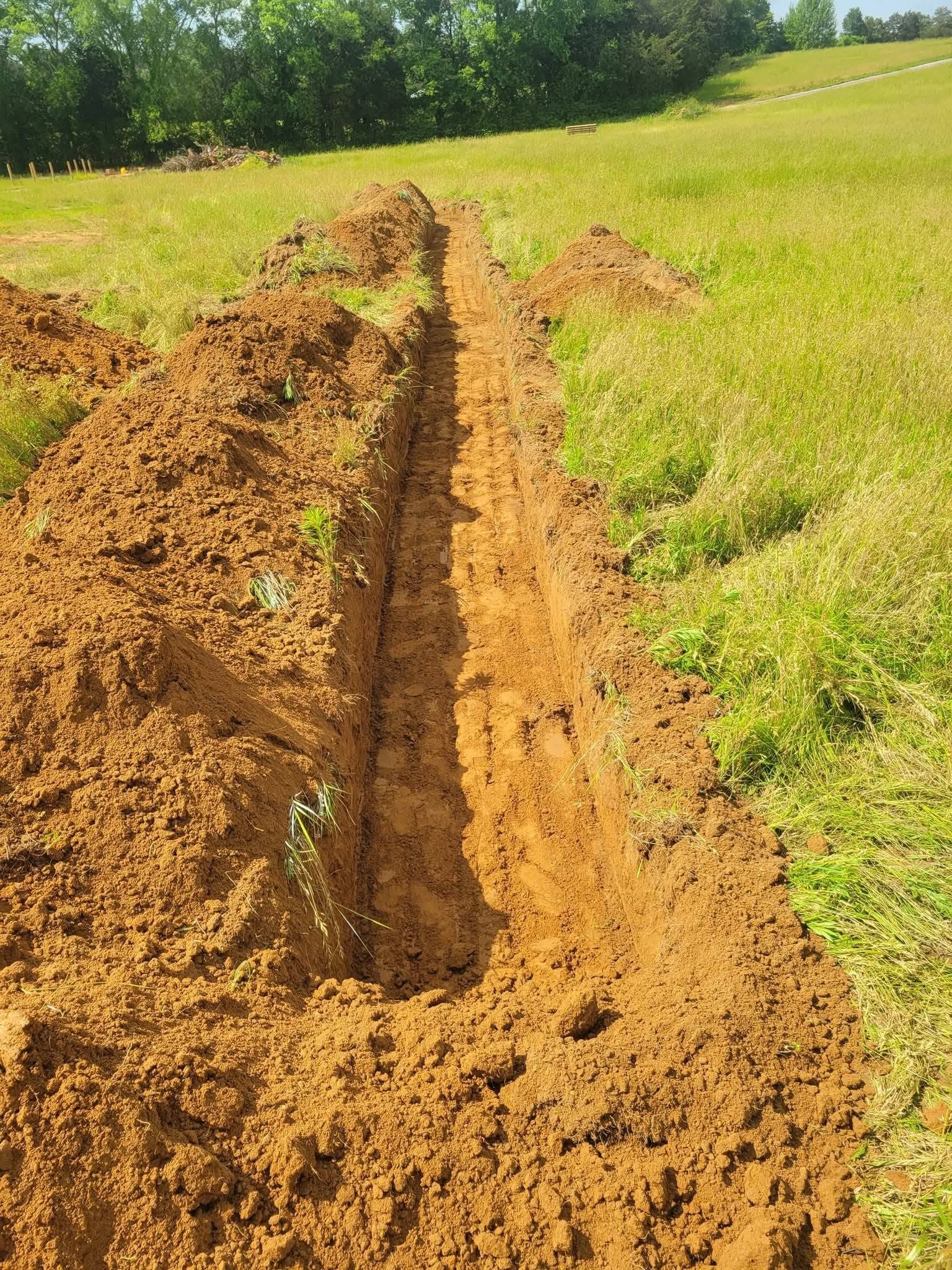 Freshly dug trench in a grassy field with a mound of dirt beside it under sunlight